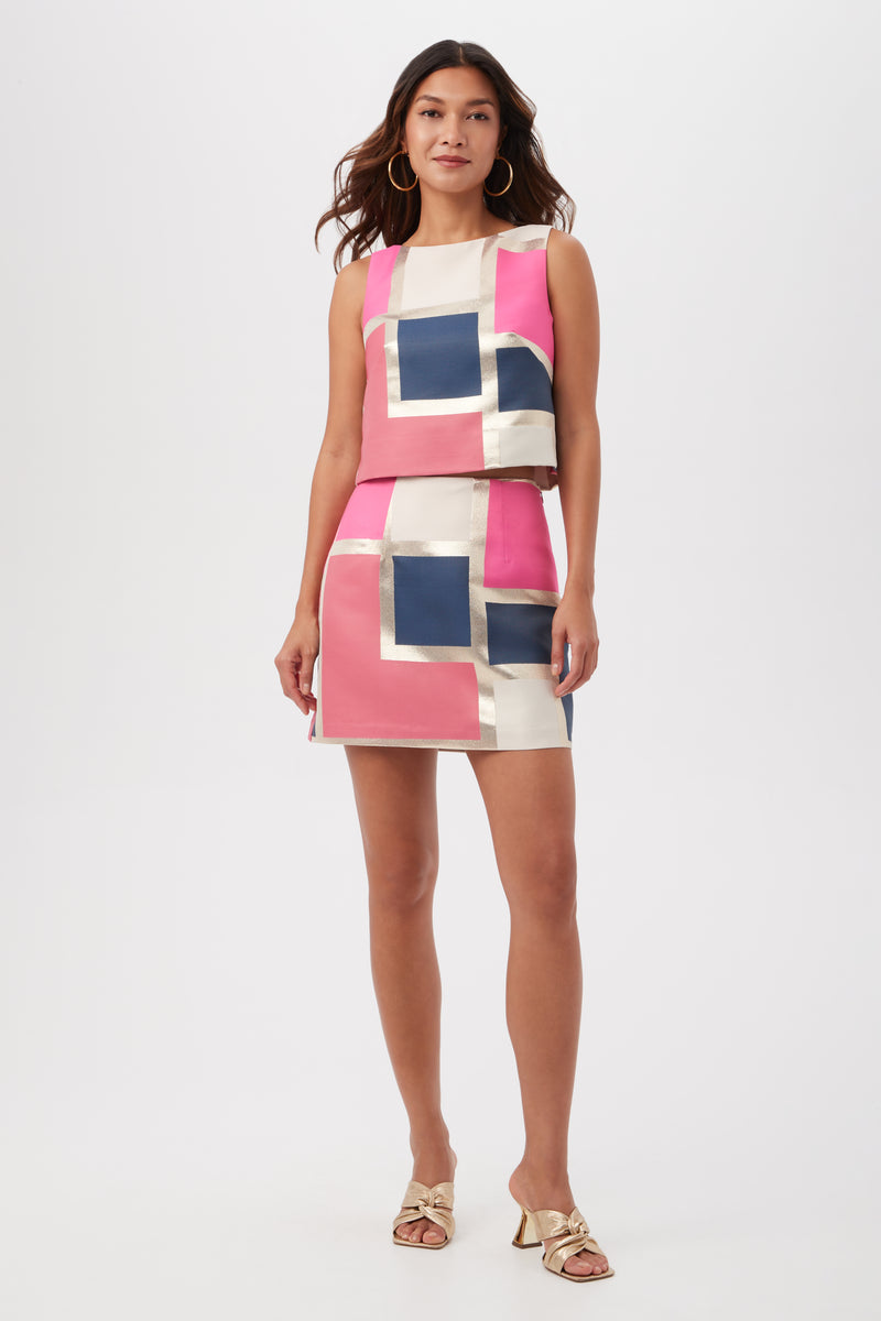 A woman stands facing forward against a plain background, wearing the High Rise Rico Skirt in Block Party Jacquard with pink, navy blue, ivory, and gold panels. She has loose wavy hair, gold hoop earrings, and gold heeled sandals.