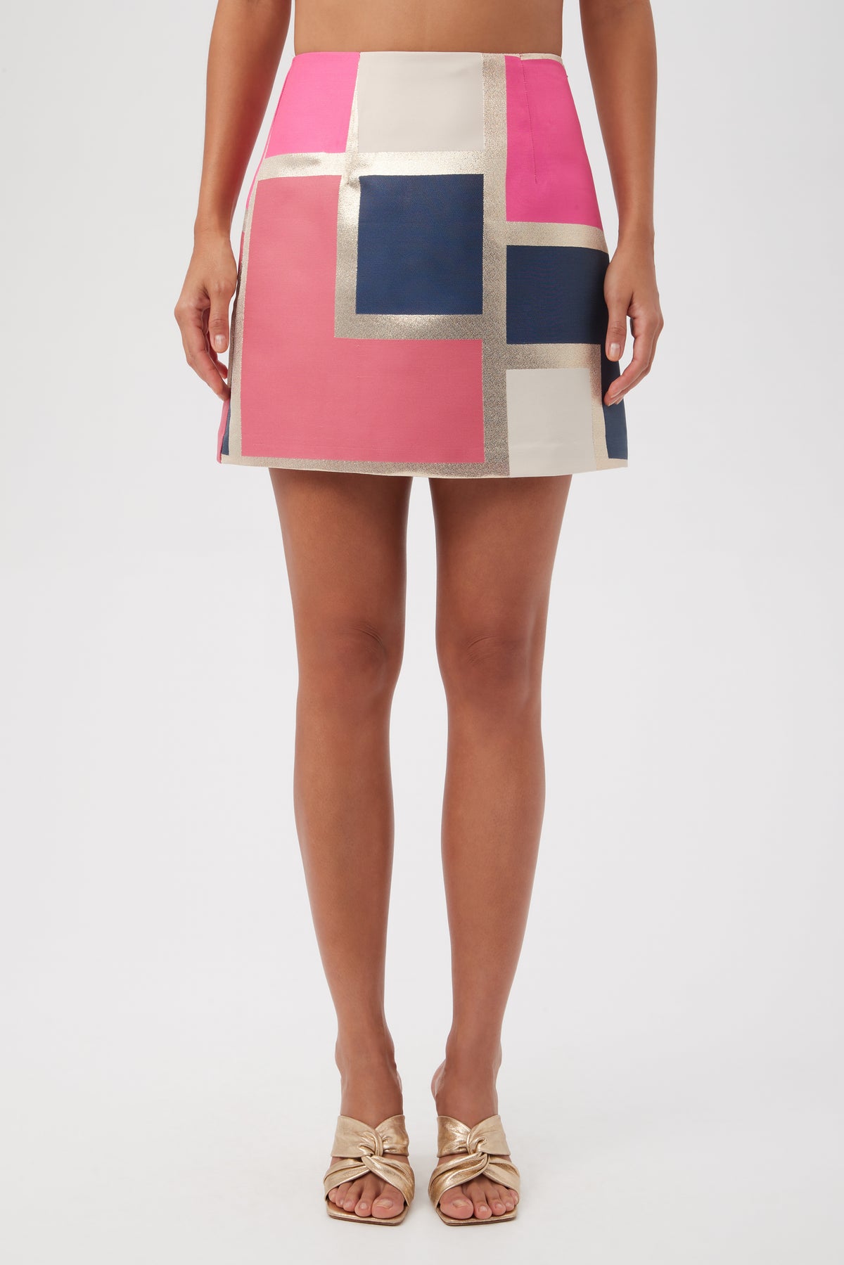 A woman wears the High Rise Rico Skirt - Block Party Jacquard in Multi, featuring geometric color blocks, paired with gold open-toe sandals. The image is cropped from midriff to feet.