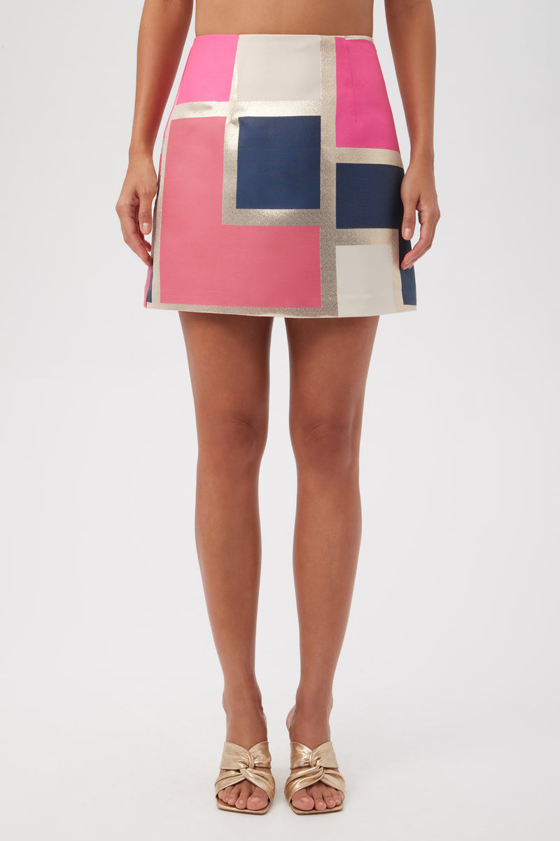 A woman wears the High Rise Rico Skirt - Block Party Jacquard in Multi, featuring geometric color blocks, paired with gold open-toe sandals. The image is cropped from midriff to feet.