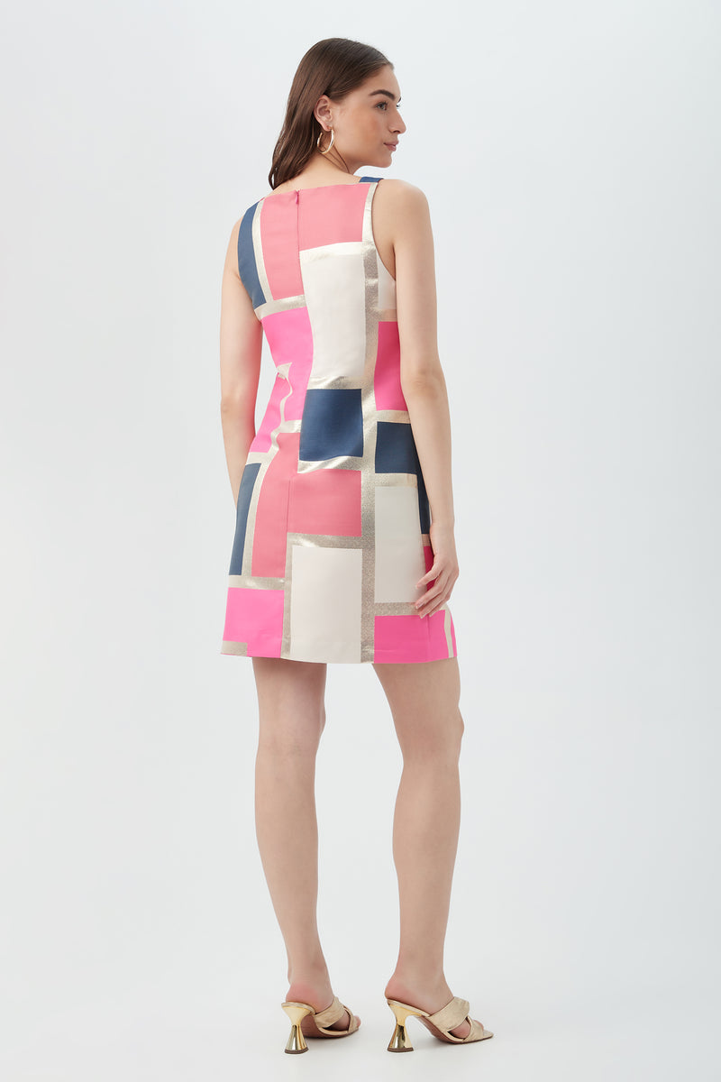 A woman with long hair faces away, wearing the Creosote Dress - Block Party Jacquard, a sleeveless, above-the-knee dress featuring a bold patchwork pattern in pink, white, navy, and beige. She pairs it with gold heeled sandals against a plain white background.
