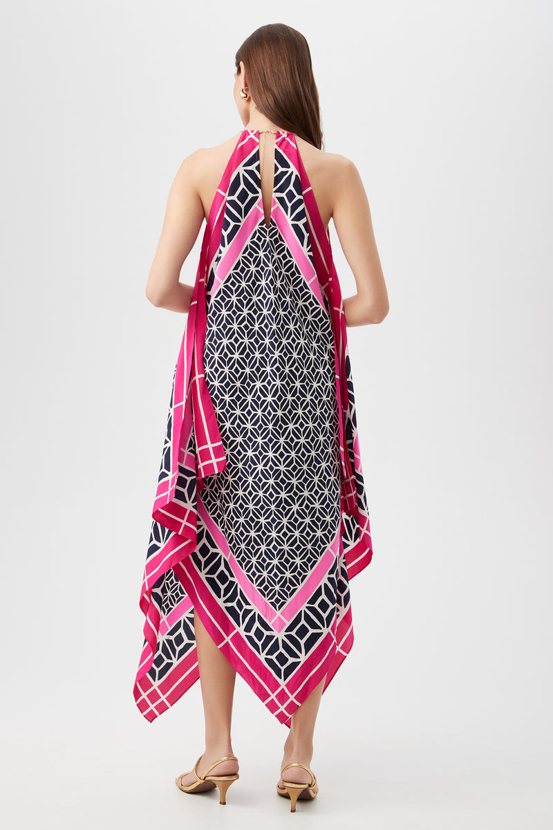 A woman with long brown hair faces away from the camera, wearing the Justinian Midi Dress - Durell Silk, a halter-neck dress featuring a geometric navy and white print with pink and white borders. She pairs it with beige heeled sandals.
