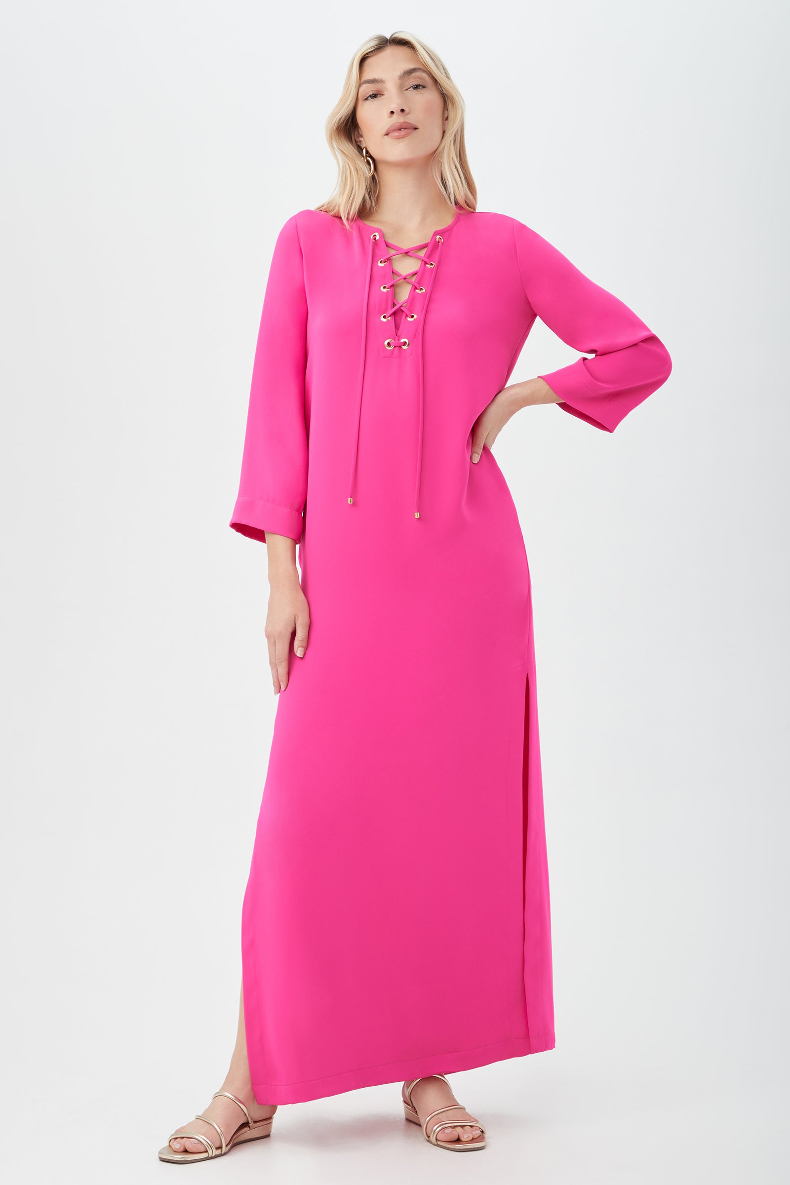 A person poses against a white background wearing the Catamaran 3/4-Sleeve Maxi Dress in Petal Pink, featuring a lace-up neckline and side slit, paired with flat sandals. Their left hand rests on their hip while their right arm is relaxed.