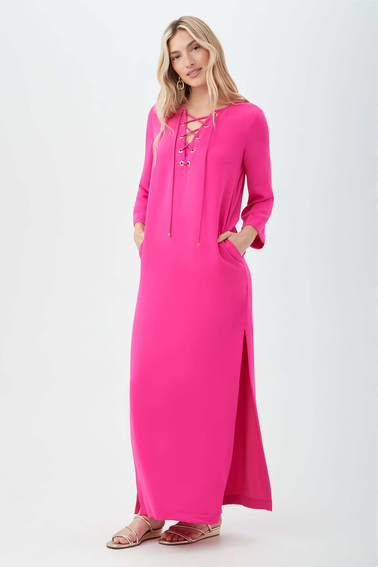 A woman with long blonde hair smiles with hands in pockets, wearing the Catamaran 3/4-Sleeve Maxi Dress—a bright pink maxi dress with a lace-up neckline and side slit—paired with flat sandals.