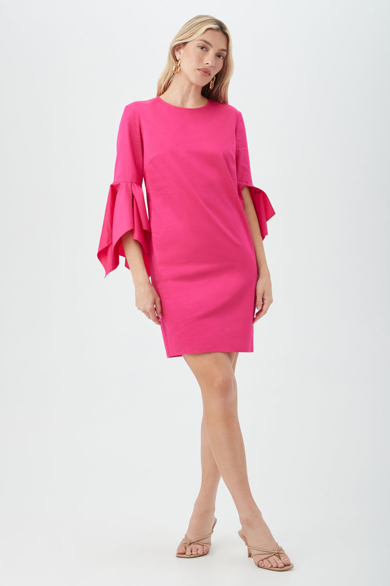 A woman models the Fontainebleau Dress in Petal Pink, a knee-length style with flared sleeves, posing against a white background with one leg crossed and wearing beige heeled sandals.