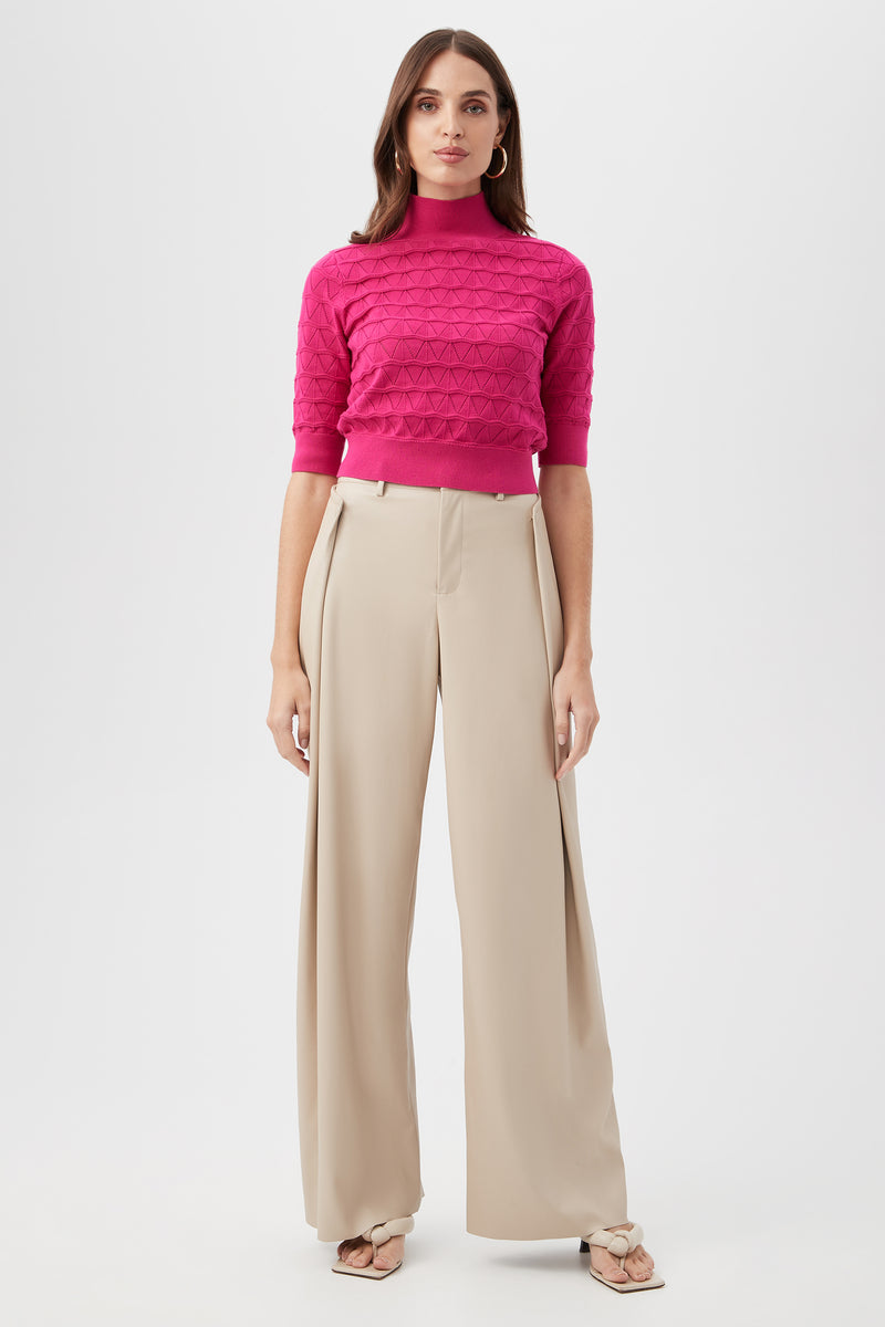 A woman stands facing forward, wearing bright pink textured short-sleeve turtleneck and high-waisted, wide-leg pleated Bexley Pant in beige with open-toe sandals. The background is plain white.