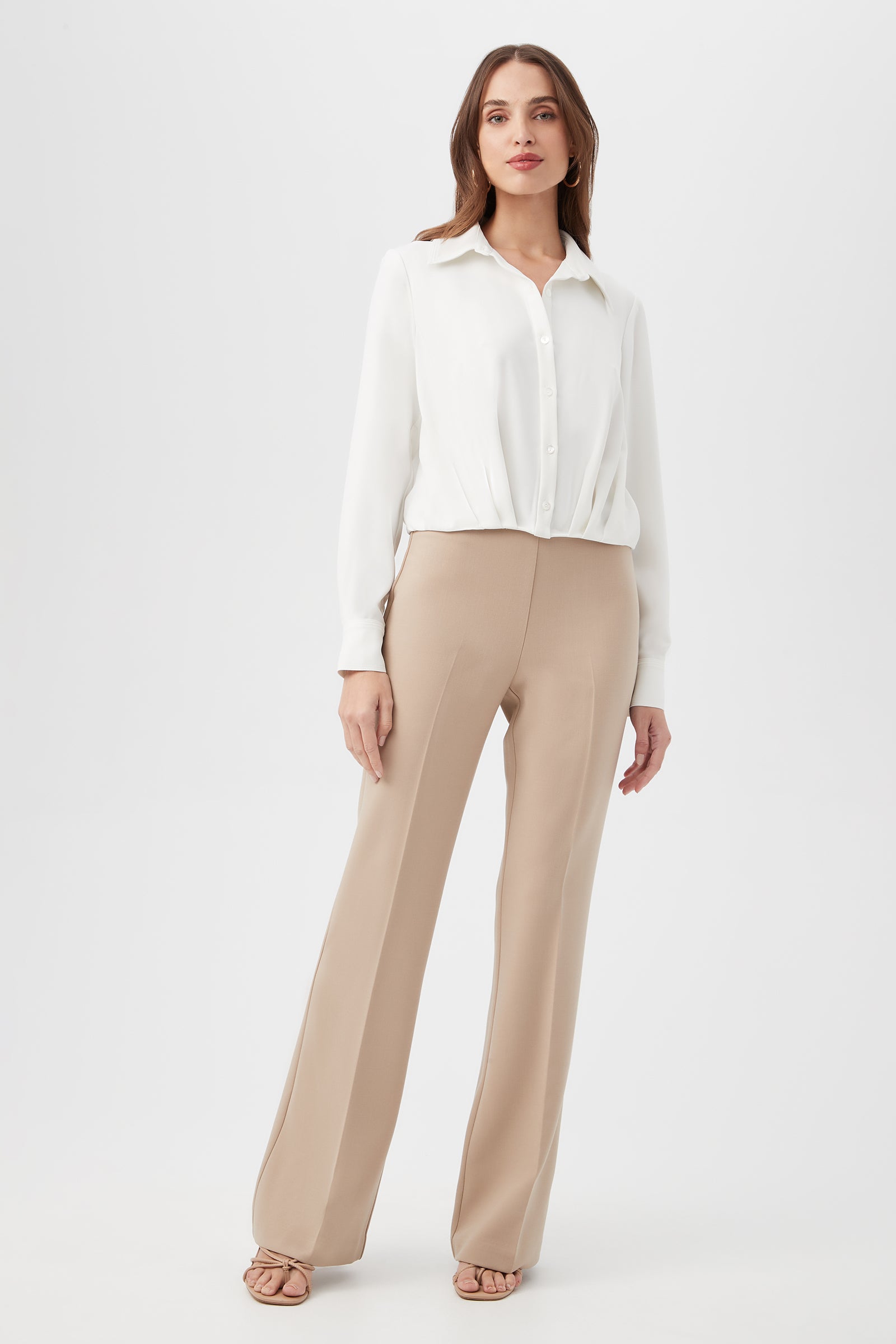 A woman stands against a white background wearing a white long-sleeve blouse tucked into high-waisted Jacoba Slim-Fit Stretch Bootcut Pants and beige open-toe heels.