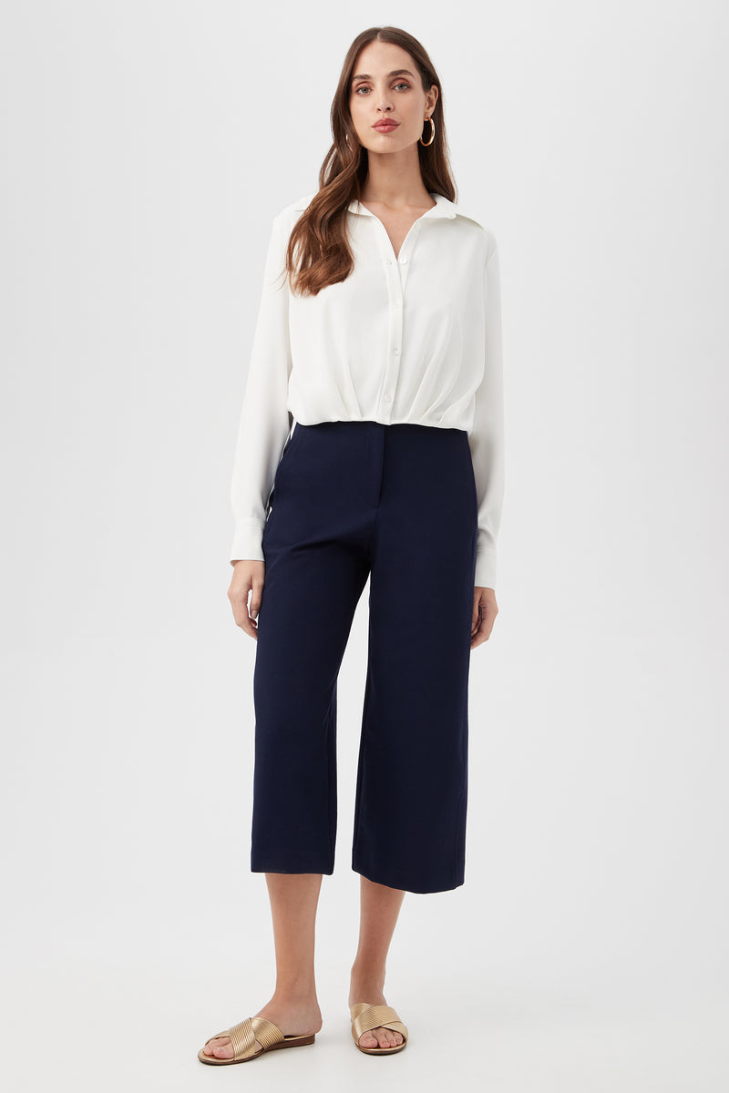 A woman with long brown hair and a neutral expression stands against a plain background, wearing a white long-sleeve blouse, beige slide sandals, and the navy blue Patyn Pant.