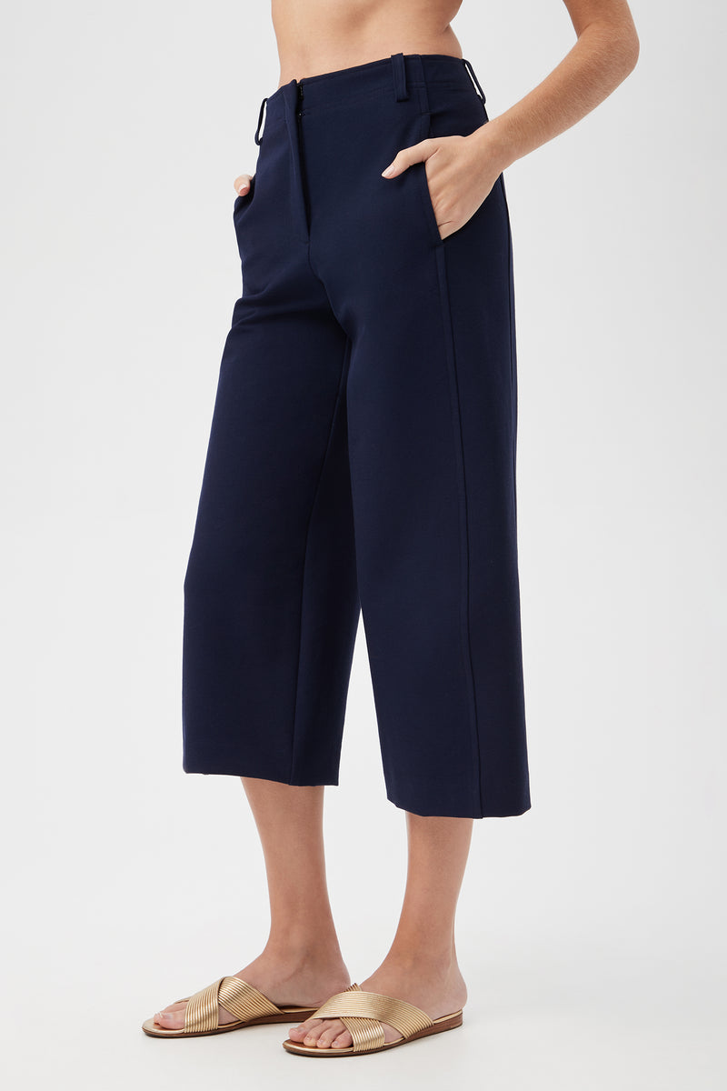 A person models the Patyn Pant—navy blue, wide-legged, and cropped—with hands in pockets and gold sandals against a plain white background.