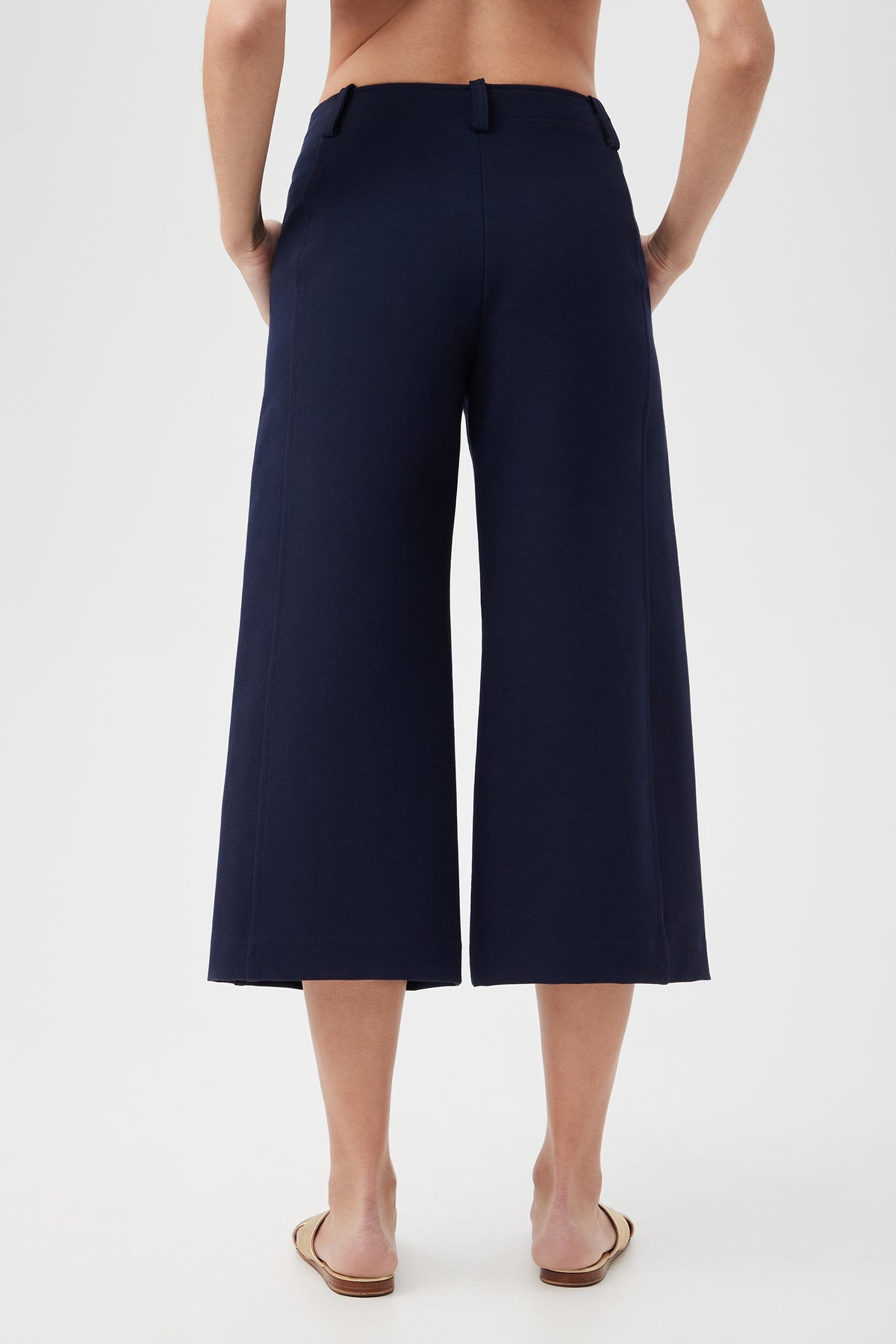 A person faces away, showcasing the Patyn Pant in navy blue—wide-leg and cropped—with tan flat shoes and a bare back above the waistband, against a plain light background.