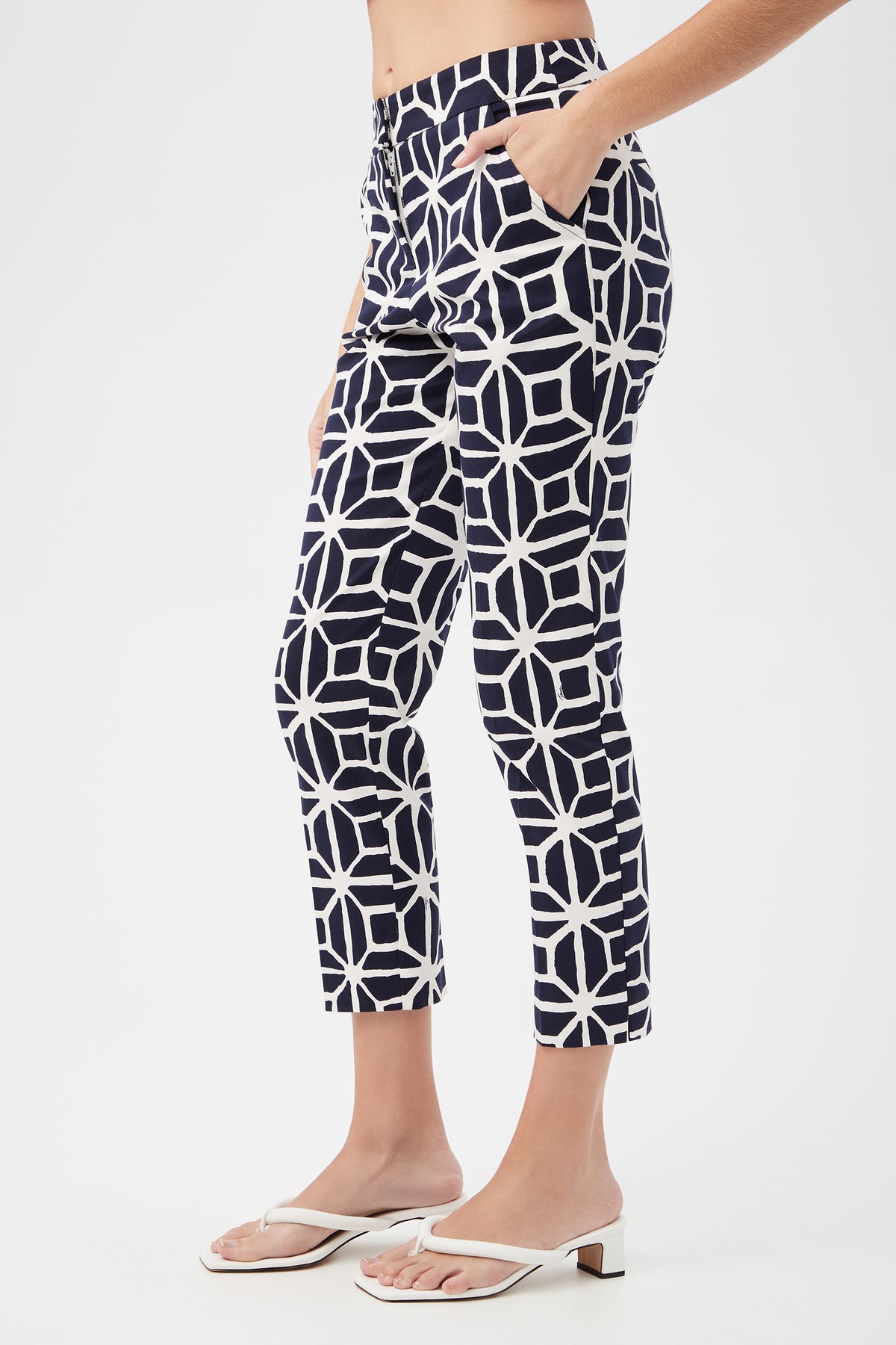 A person wearing the Moss 2 Slim Crop Pant - Durell Herringbone and white heeled sandals, shown from the side with one hand in a pocket against a plain background.