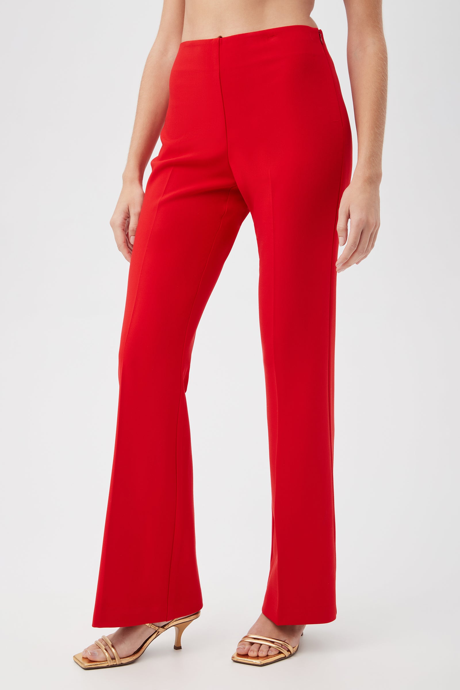 A person shown from mid-torso down wears Jacoba Slim-Fit Stretch Bootcut Pants in Reina Red and gold heeled sandals, set against a plain light background.