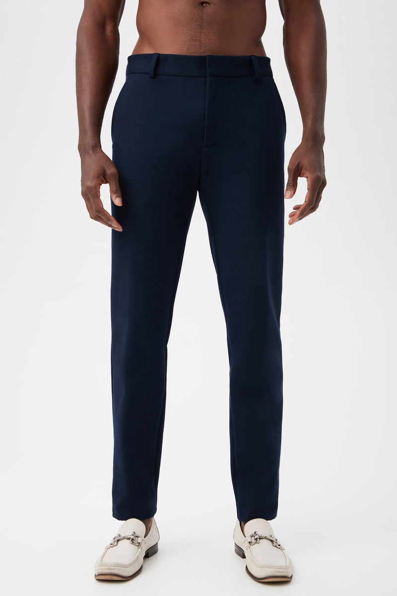 A man models the Clyde Slim Trouser - Stretch PiquÃ© in indigo with white loafers, standing against a plain white background; his torso is bare and arms relaxed at his sides.