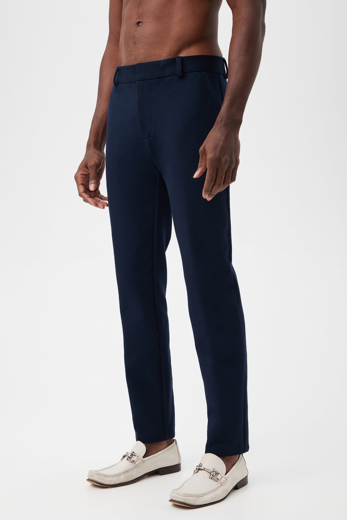 A shirtless man models the Clyde Slim Trouser - Stretch PiquÃ© in navy blue and white loafers, standing against a plain white background.