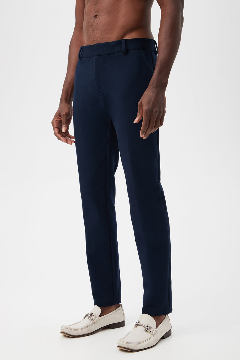 A shirtless man models the Clyde Slim Trouser - Stretch PiquÃ© in navy blue and white loafers, standing against a plain white background.