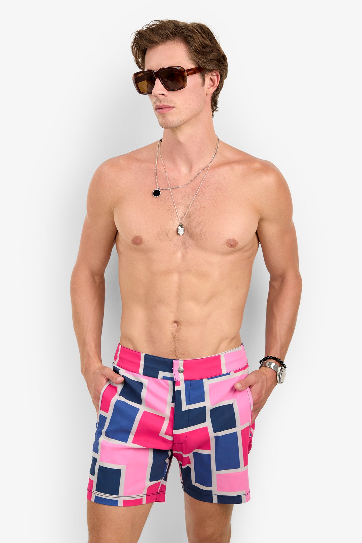 Shirtless man in sunglasses, layered necklaces, watch, and the Honolulu Boardshort—pink swim trunks with a geometric blue and white pattern—standing against a plain light background.