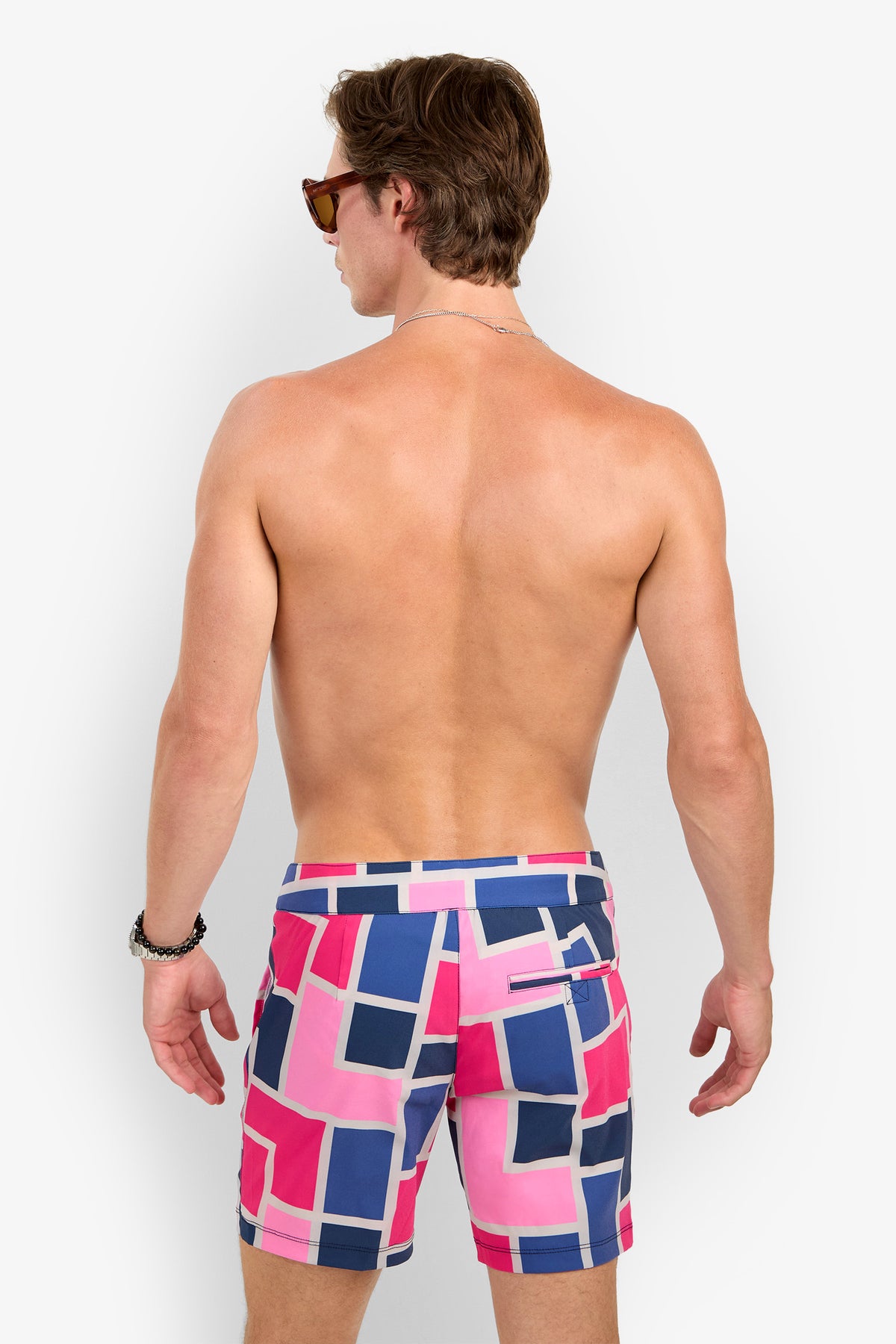 A man with short brown hair wears sunglasses and the Honolulu Boardshort—geometric pink, blue, and white swim trunks. He is shirtless, facing away from the camera against a plain white background.