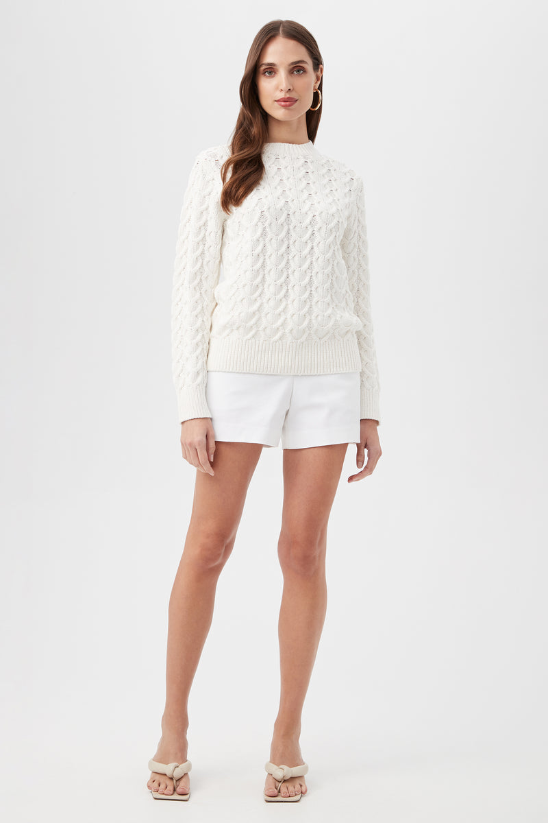 A woman with long brown hair and a neutral expression stands against a white background, wearing the Corazon Heart Knit Crewneck Sweater, white shorts, and beige heeled sandals.