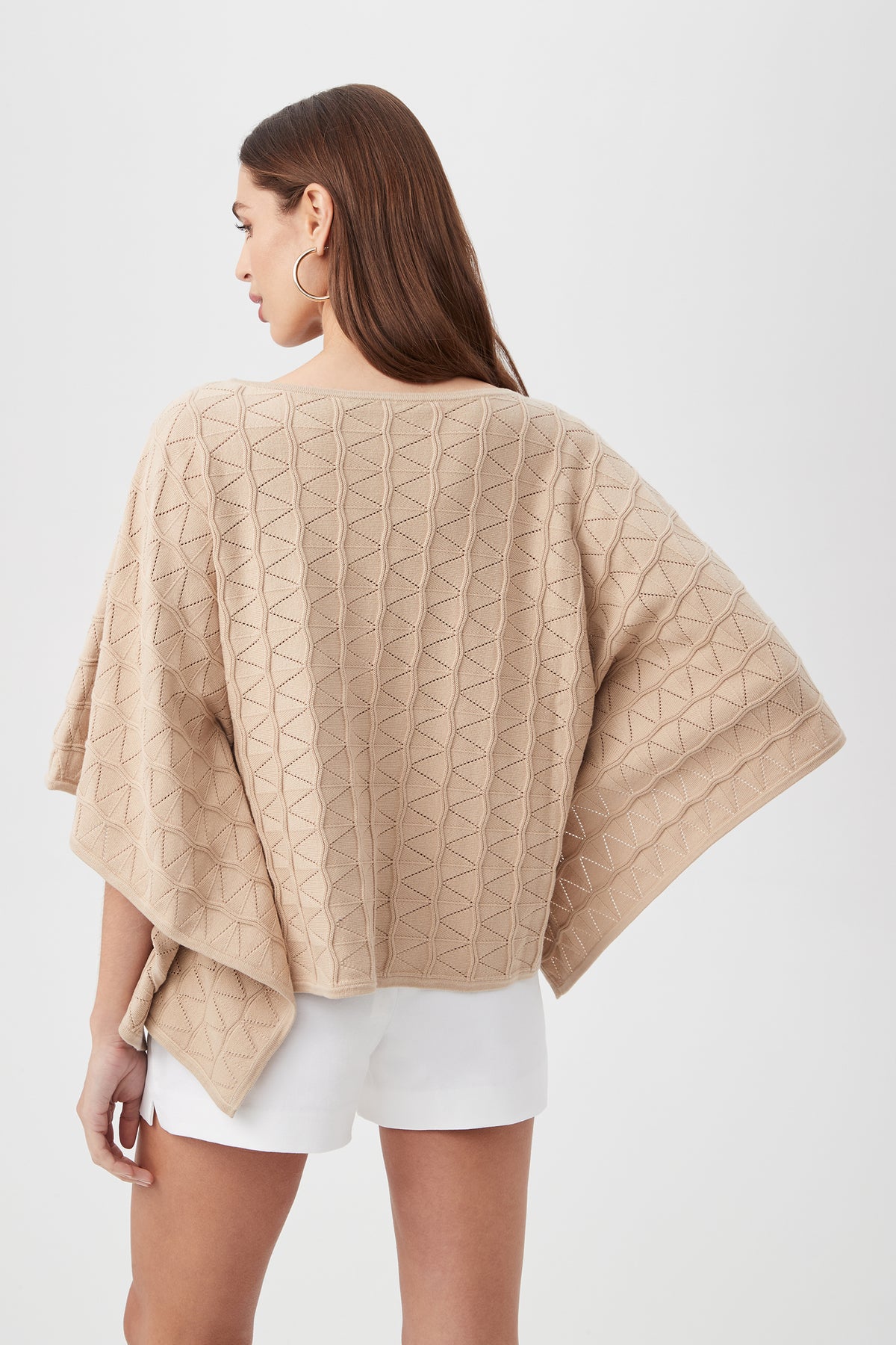 A woman with long brown hair, large hoop earrings, and white shorts models the Chic Wrap 2 Sweater—a beige textured poncho with a geometric pattern—while standing against a plain white background.