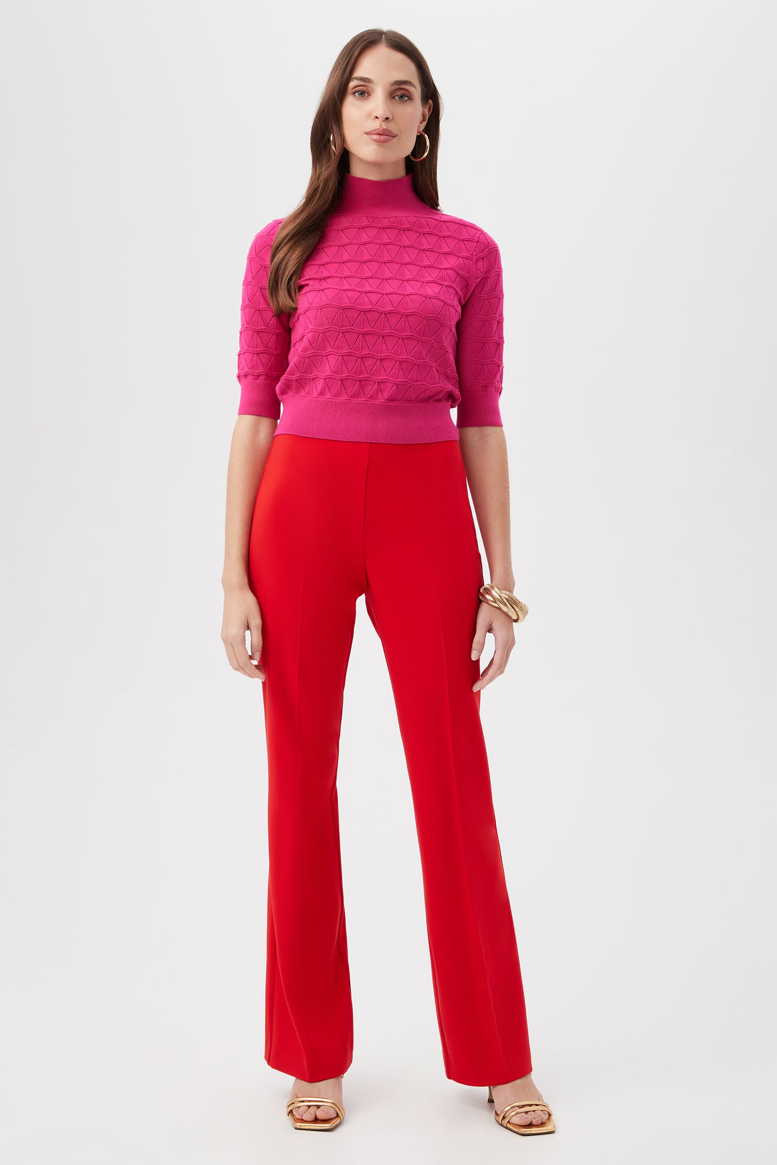 A woman stands against a white background, wearing a textured pink short-sleeve turtleneck sweater, Jacoba Slim-Fit Stretch Bootcut Pants in red, gold sandals, hoop earrings, chunky bracelets, with straight hair and a neutral expression.