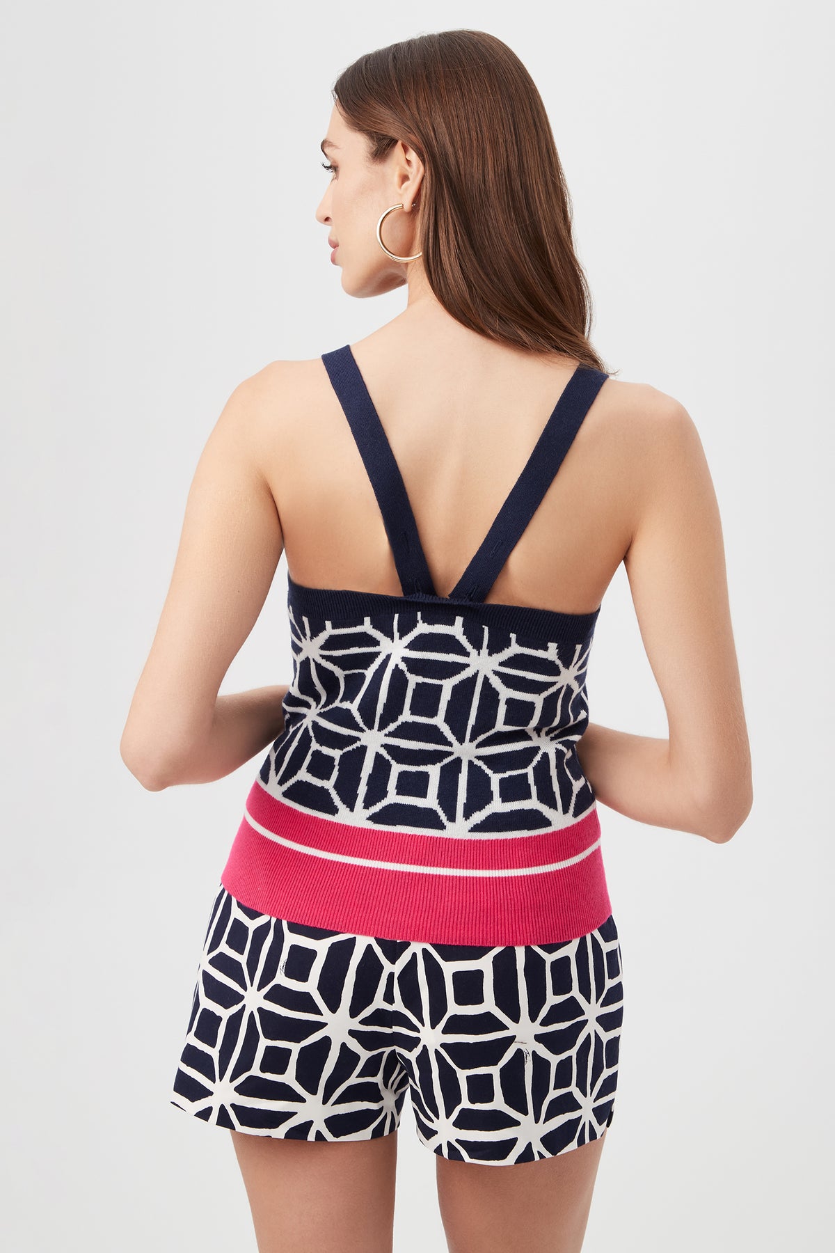 A woman with long brown hair, shown from the back against a plain light background, wears the Casady Sleeveless Cami Top - Durell Knit paired with matching geometric navy and white shorts featuring a wide pink waistband.