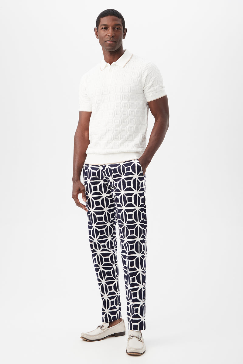 A man stands against a white background wearing the Camilo Short-Sleeve Polo in white, paired with navy pants featuring a bold white geometric pattern and light-colored shoes.