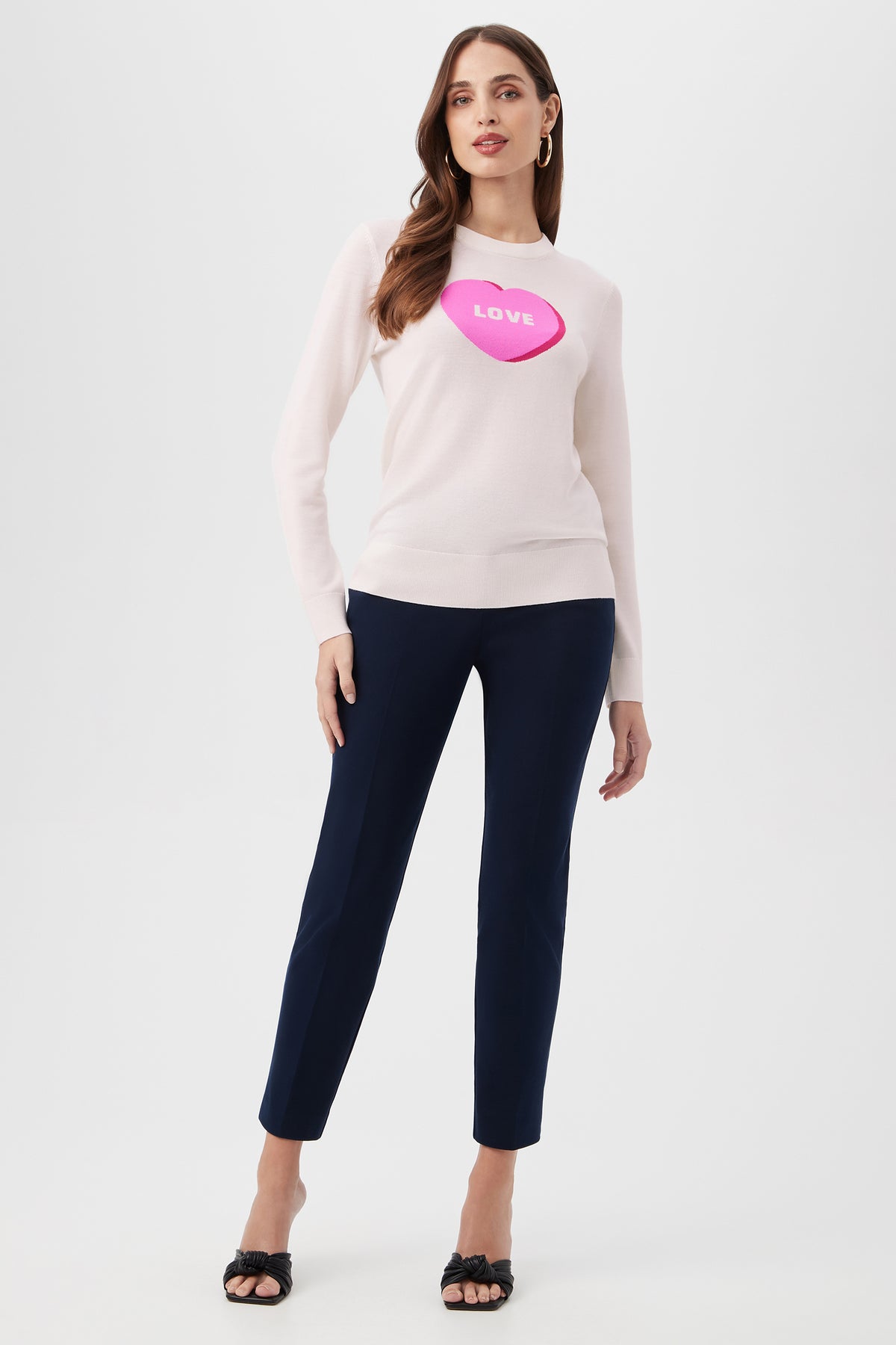 A woman wears the Love Merino Wool Crewneck Sweater in light pink with a large pink candy heart labeled LOVE, paired with navy ankle-length pants and black open-toe heels, standing against a plain white background.