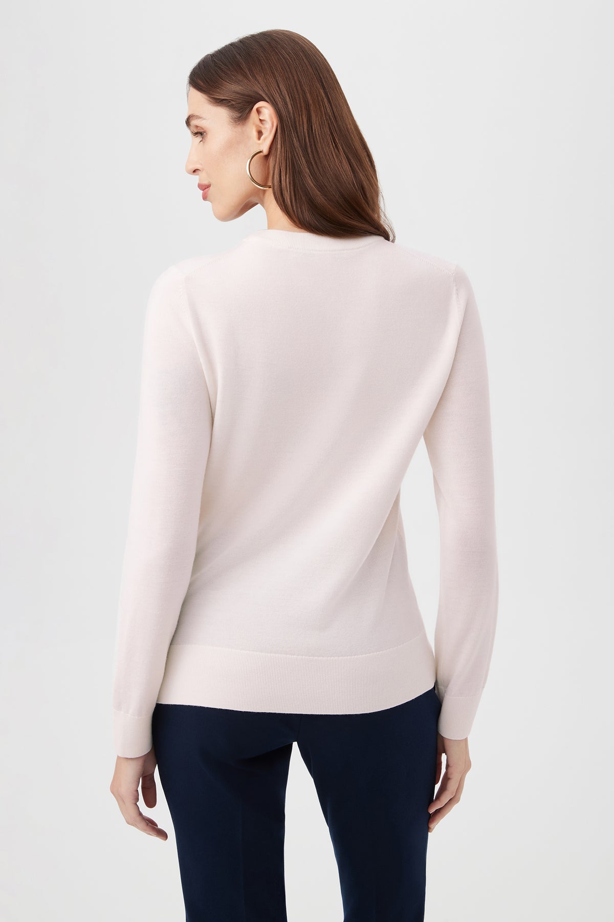 A woman with long brown hair wears the Love Merino Wool Crewneck Sweater in light pink and dark pants, standing with her back to the camera against a plain light background.