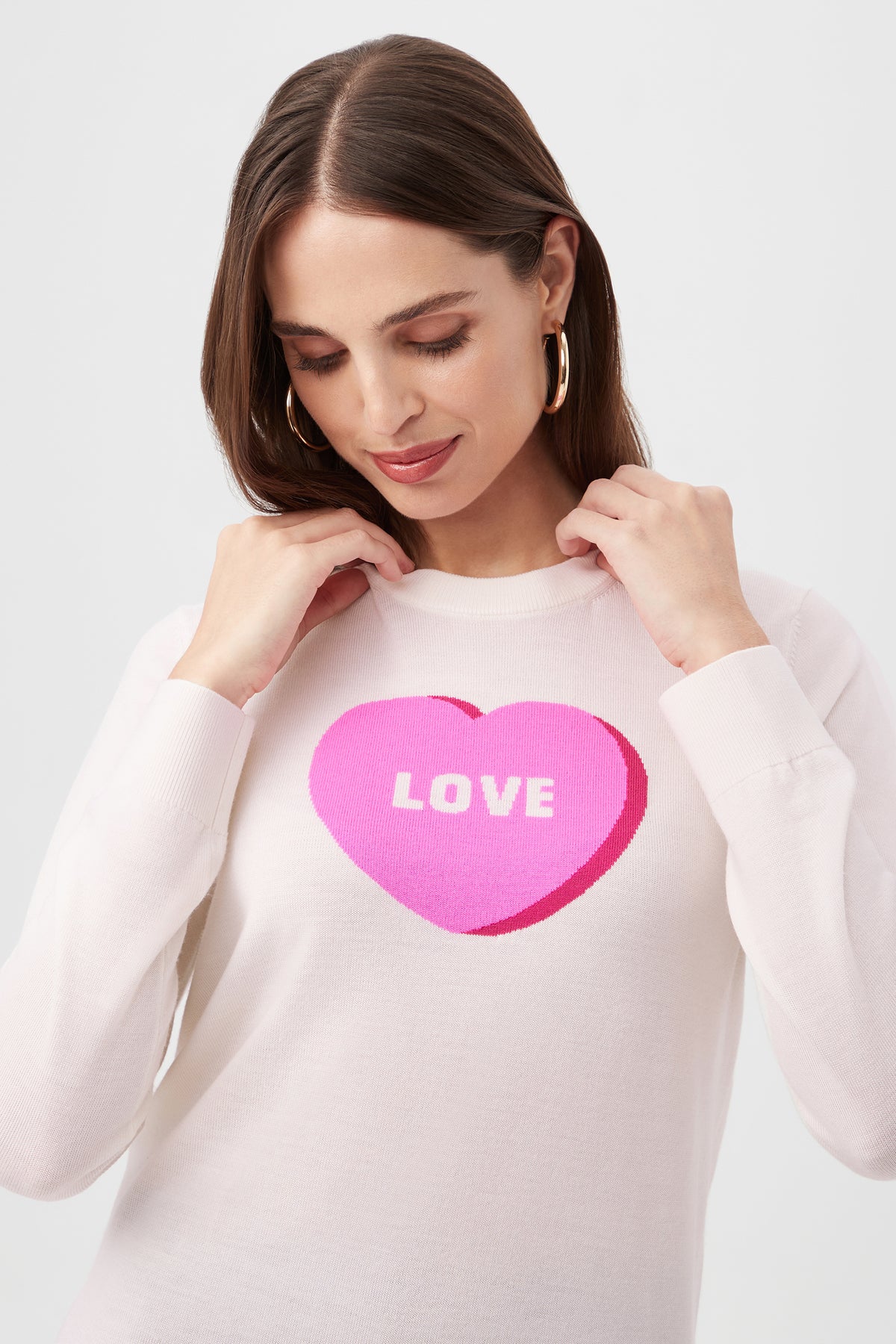 A woman with brown hair and gold hoop earrings wears the Love Merino Wool Crewneck Sweater, a white top featuring a large pink heart with LOVE inside. She looks down, gently holding the collar of her sweater.