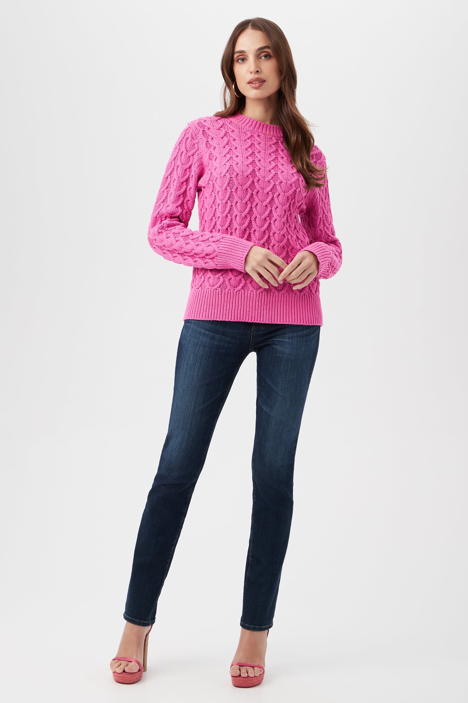 A woman with long brown hair and a neutral expression stands against a white background, wearing the Corazon Heart Knit Crewneck Sweater in bright pink, dark blue jeans, and pink high-heeled sandals.