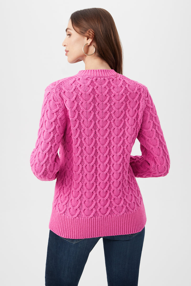 A woman with long brown hair models the Corazon Heart Knit Crewneck Sweater in bright pink, paired with dark jeans. Facing away from the camera, she highlights the sweater’s textured back and wears large hoop earrings.