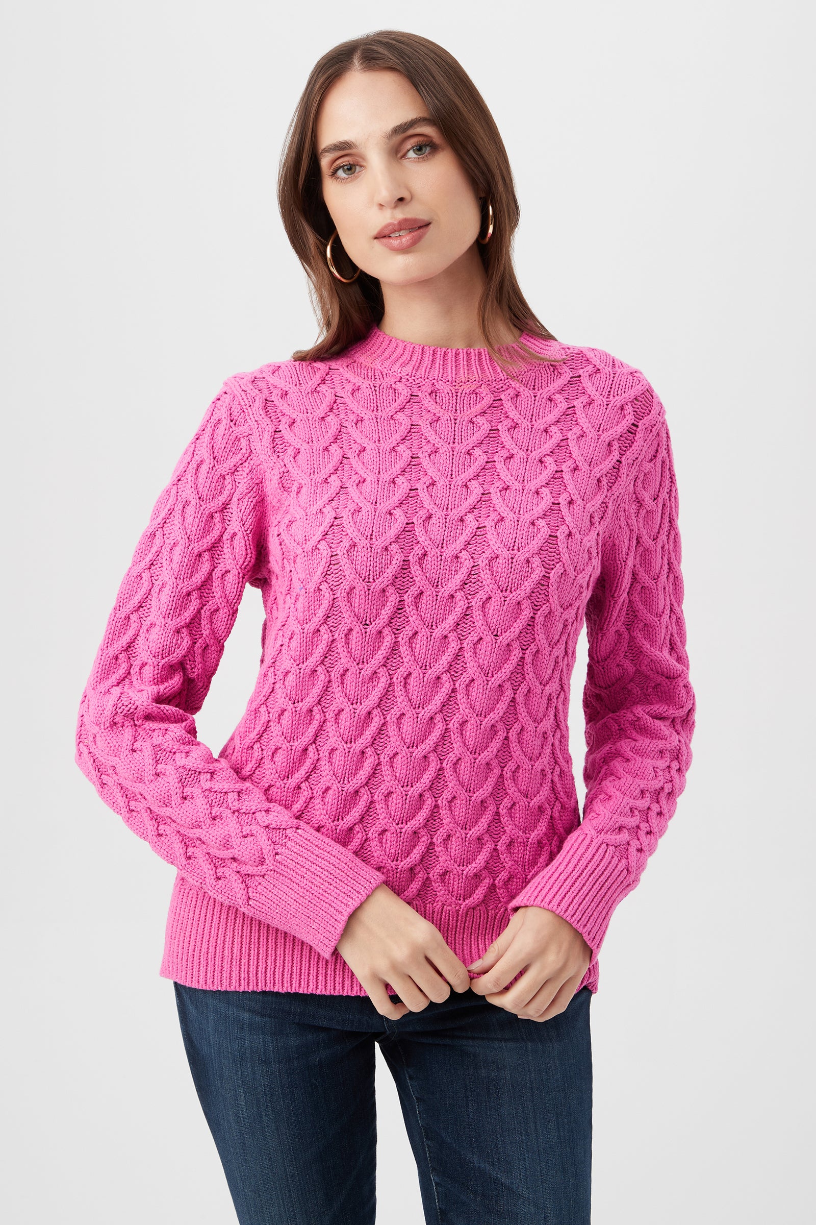 A woman wearing the Corazon Heart Knit Crewneck Sweater in Snapdragon and blue jeans stands against a plain light background, looking at the camera with a neutral expression.