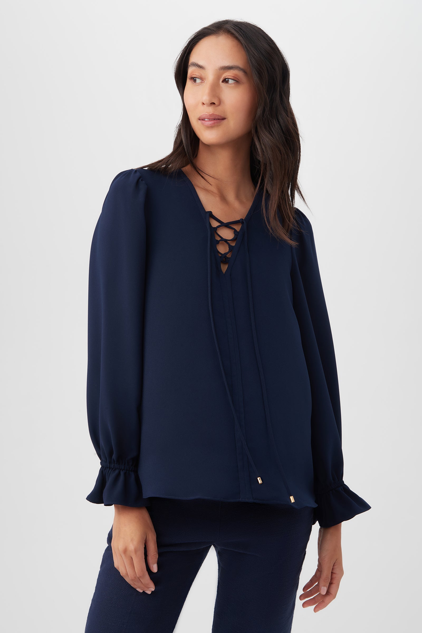 A woman with long dark hair wears the Zahara Top in indigo, featuring a lace-up V-neck and ruffled cuffs, paired with dark pants and standing against a plain light background.