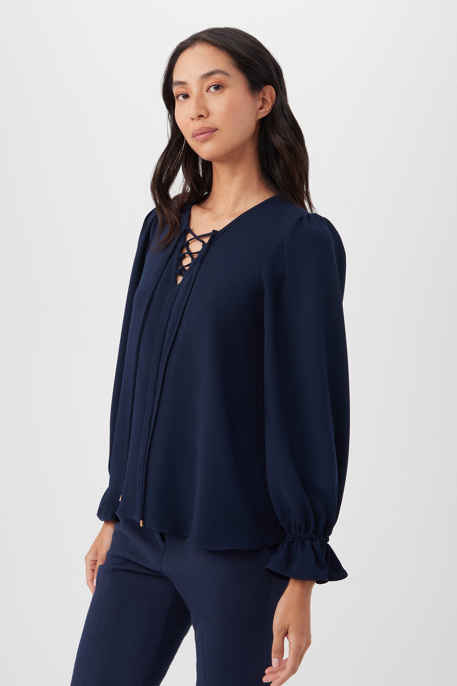 A woman wears the Zahara Top—a navy satin-back crepe long sleeve with a lace-up neckline and ruffled cuffs—paired with matching recycled polyester pants, standing against a plain light background.