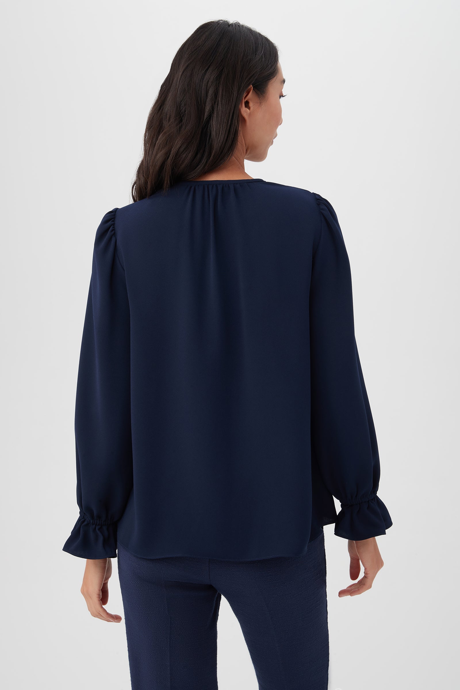 A woman with wavy dark hair is seen from the back, wearing the navy blue Zahara Top with long balloon sleeves and matching pants, standing against a plain light background.