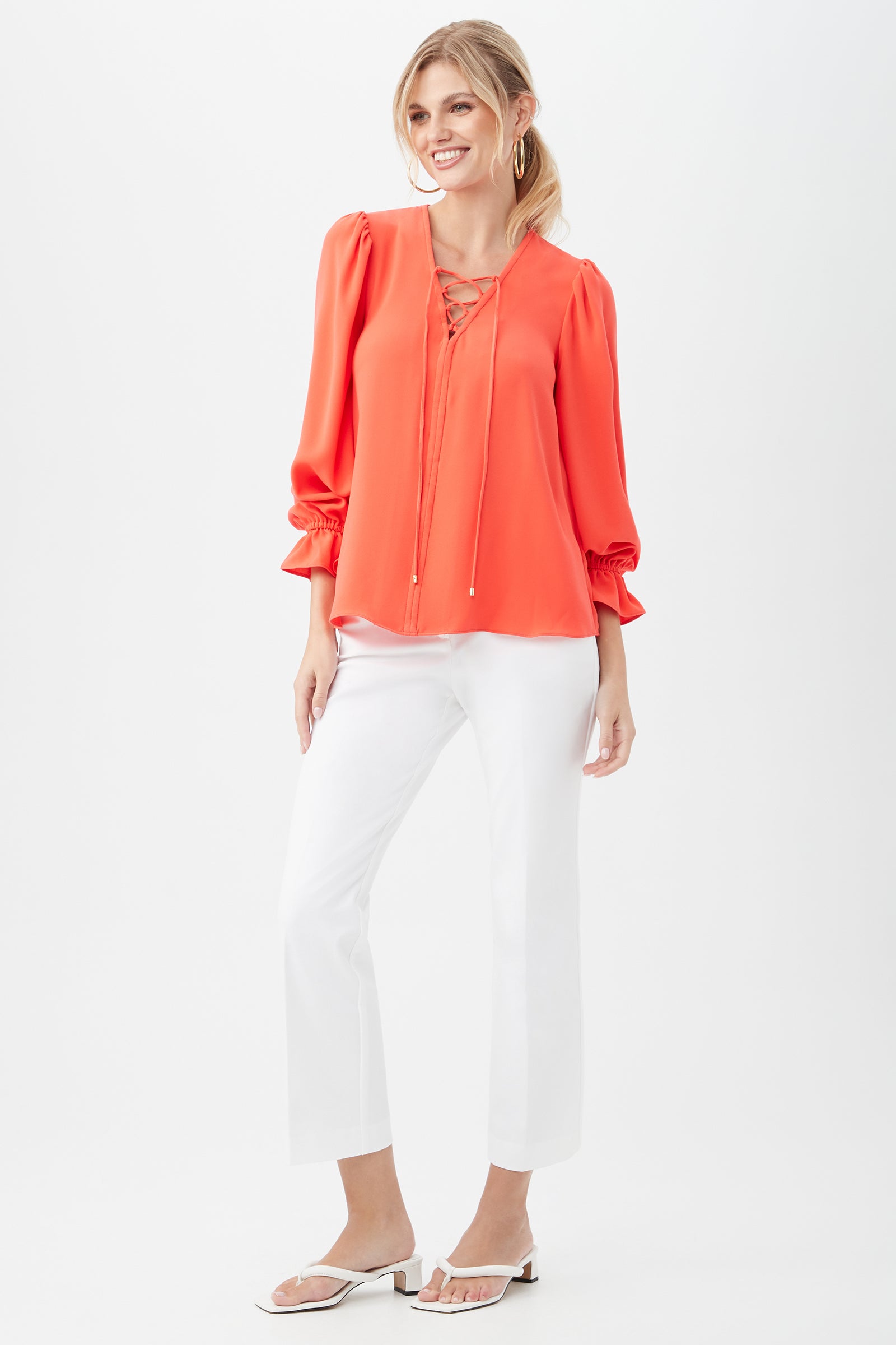 A woman smiles in the Zahara Top, a bright coral long-sleeve made from recycled polyester satin-back crepe. She pairs it with white pants and open-toe sandals, standing against a plain white background.