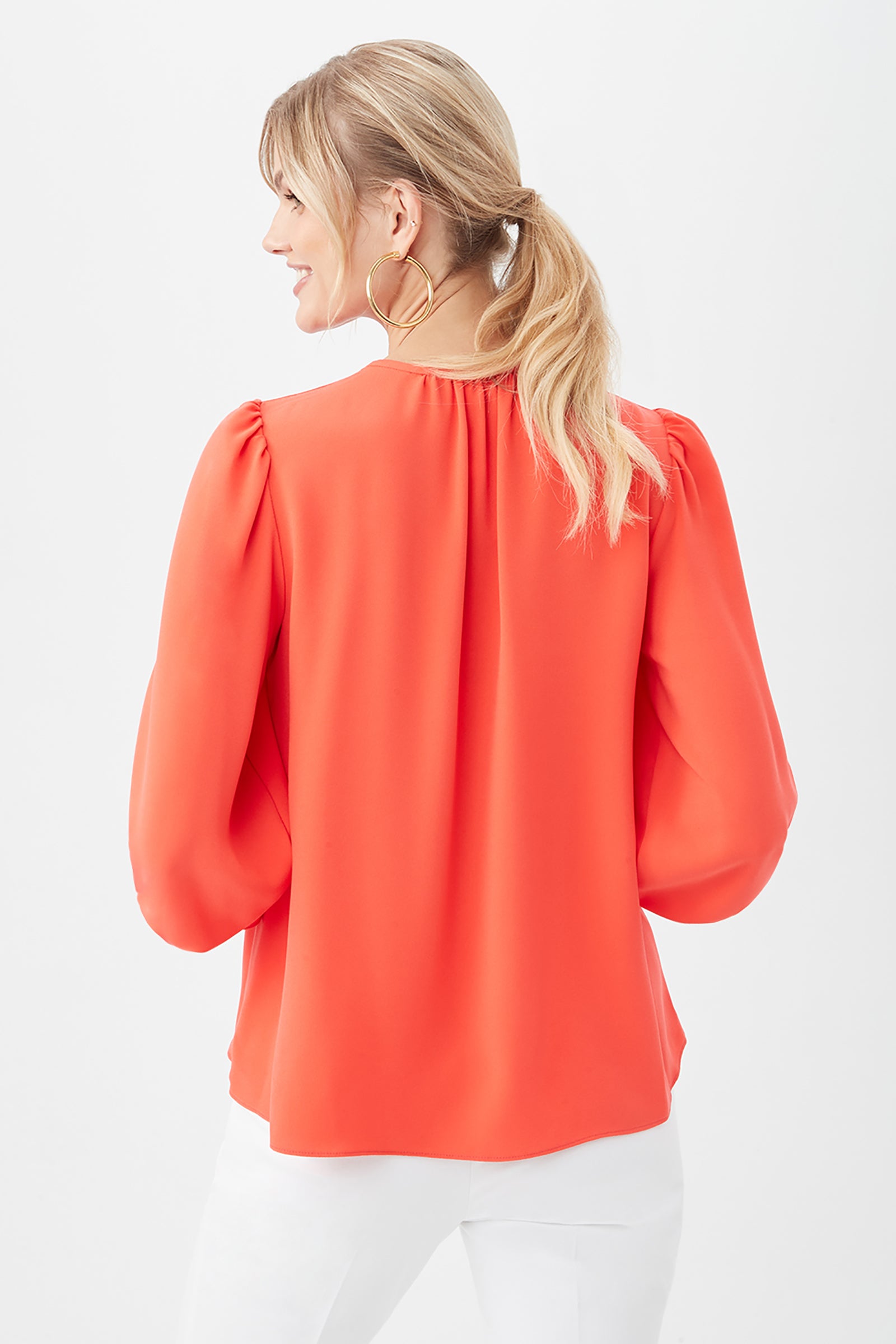 A woman with blonde hair in a ponytail wears the Zahara Top, a bright orange long-sleeve blouse, paired with white pants. She stands smiling with her back to the camera against a plain white background.