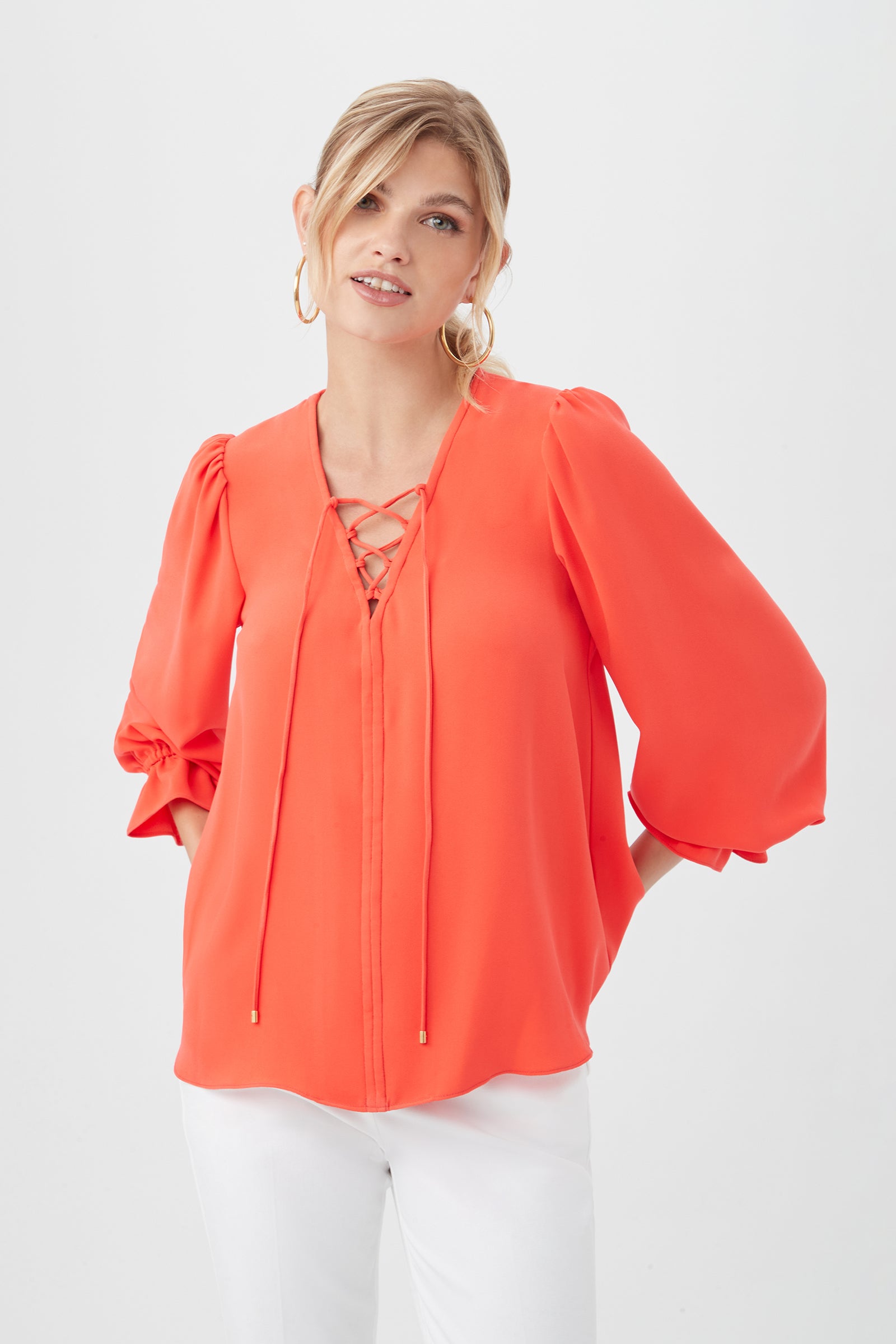 A woman wears the Zahara Top in poppy, a dressy long-sleeve blouse with puffed sleeves and a lace-up neckline made from recycled polyester, paired with white pants against a plain white background.