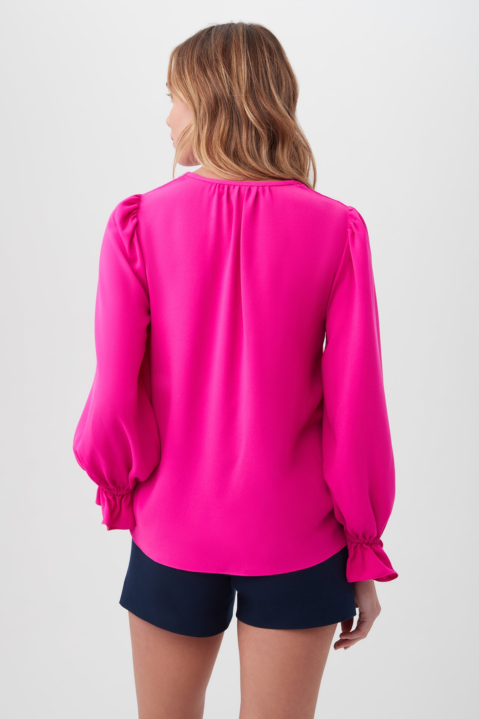 A woman with wavy light brown hair stands with her back to the camera, wearing the bright pink Zahara Top, made from recycled polyester satin-back crepe with long balloon sleeves, and dark navy shorts against a plain light background.