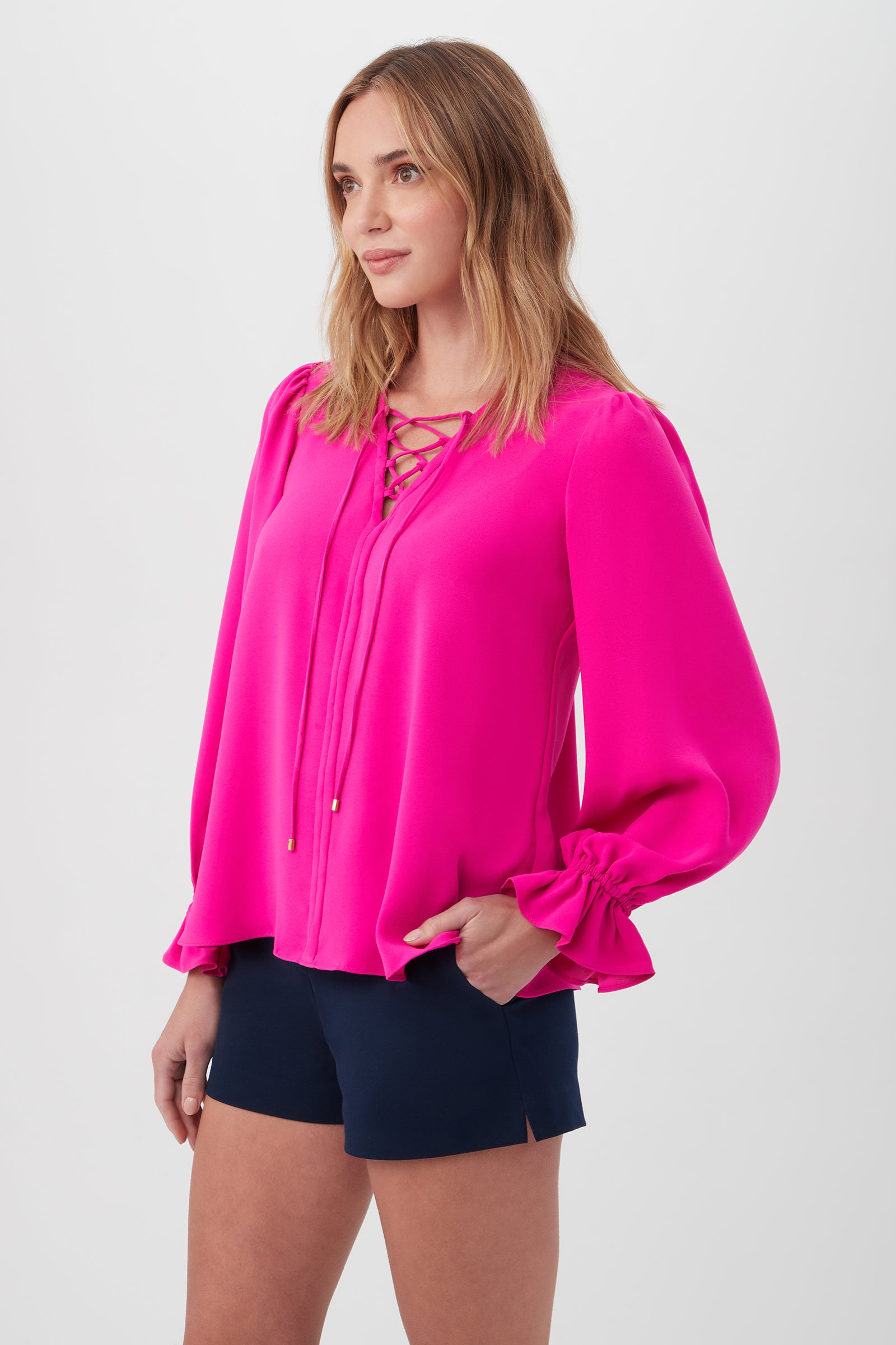 A woman wears the Zahara Top—a bright pink recycled polyester blouse with a lace-up neckline—and navy blue shorts, standing against a plain white background with one hand in her pocket, looking slightly to the side.