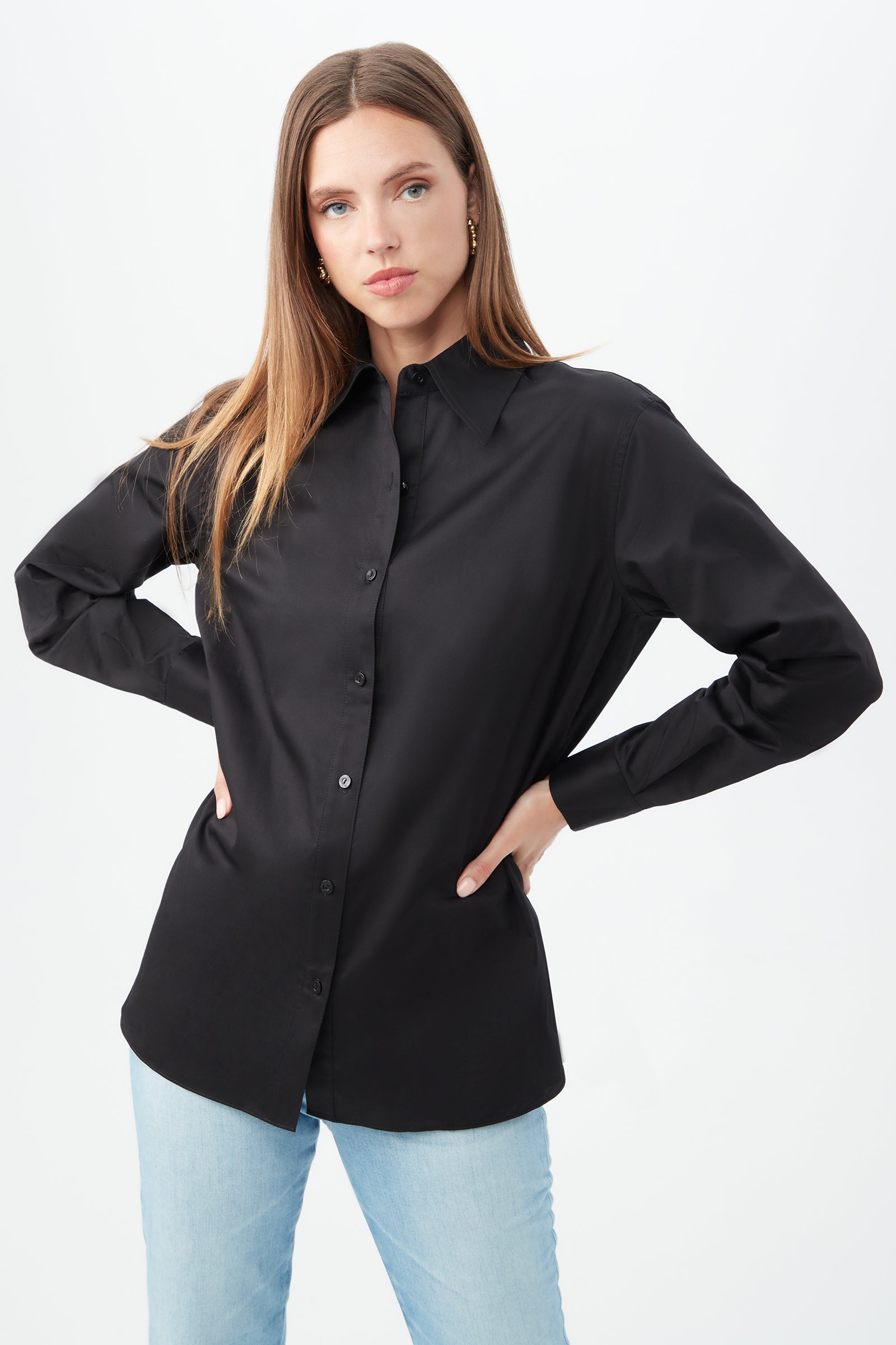 A woman with long light brown hair models the black Litsa Button-Up Cotton Shirt, paired with light blue jeans, posing with one hand on her hip against a plain white background.