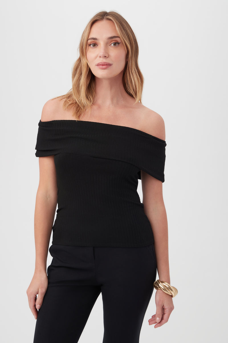 A woman with shoulder-length blonde hair, wearing the black Prizma Off-Shoulder Top and black pants, stands against a plain light background. She has a neutral expression and a gold bangle on her left wrist.