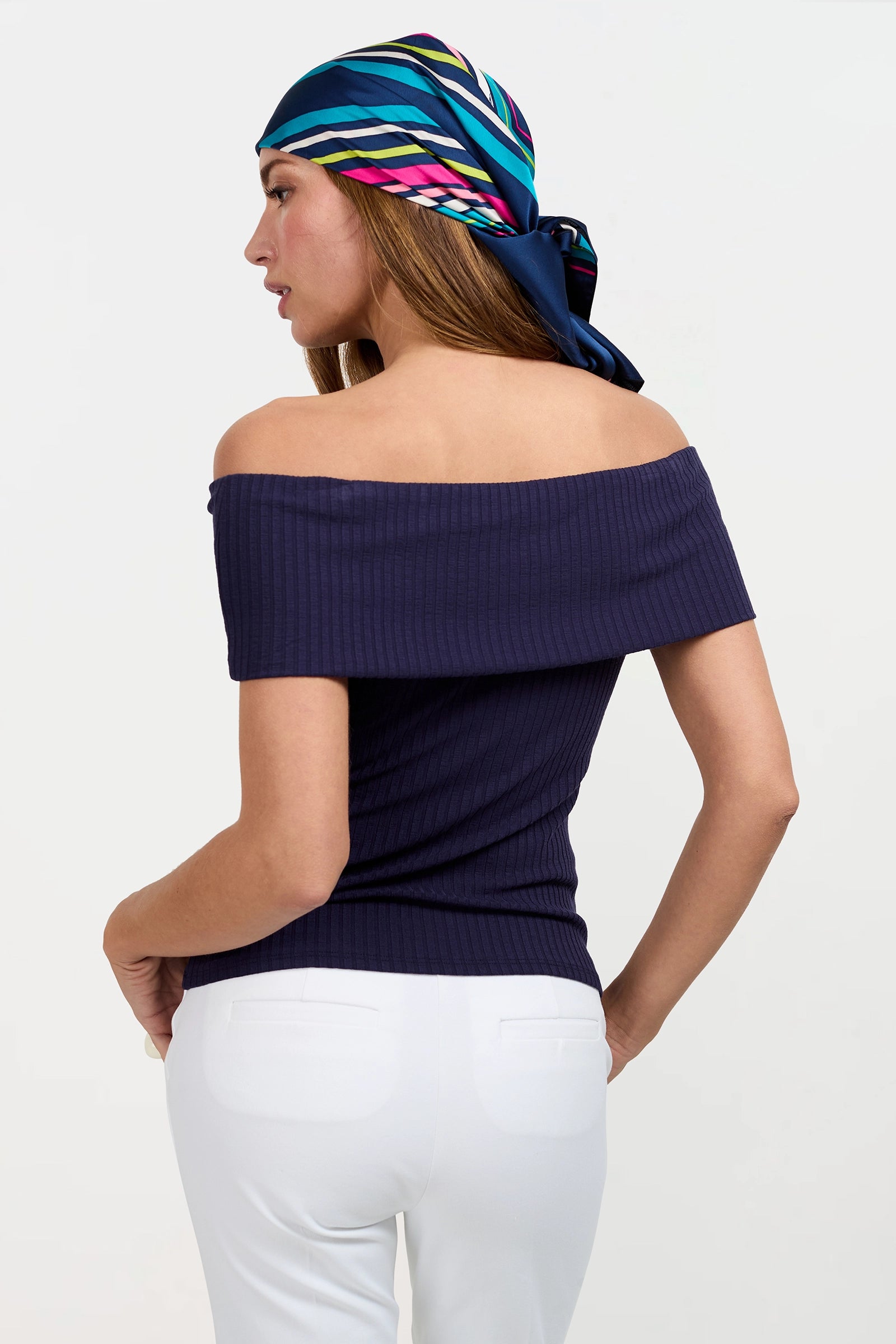A woman wearing the Prizma Off-Shoulder Top in dark navy, paired with a colorful striped headscarf and white pants, stands with her back slightly turned toward the camera.