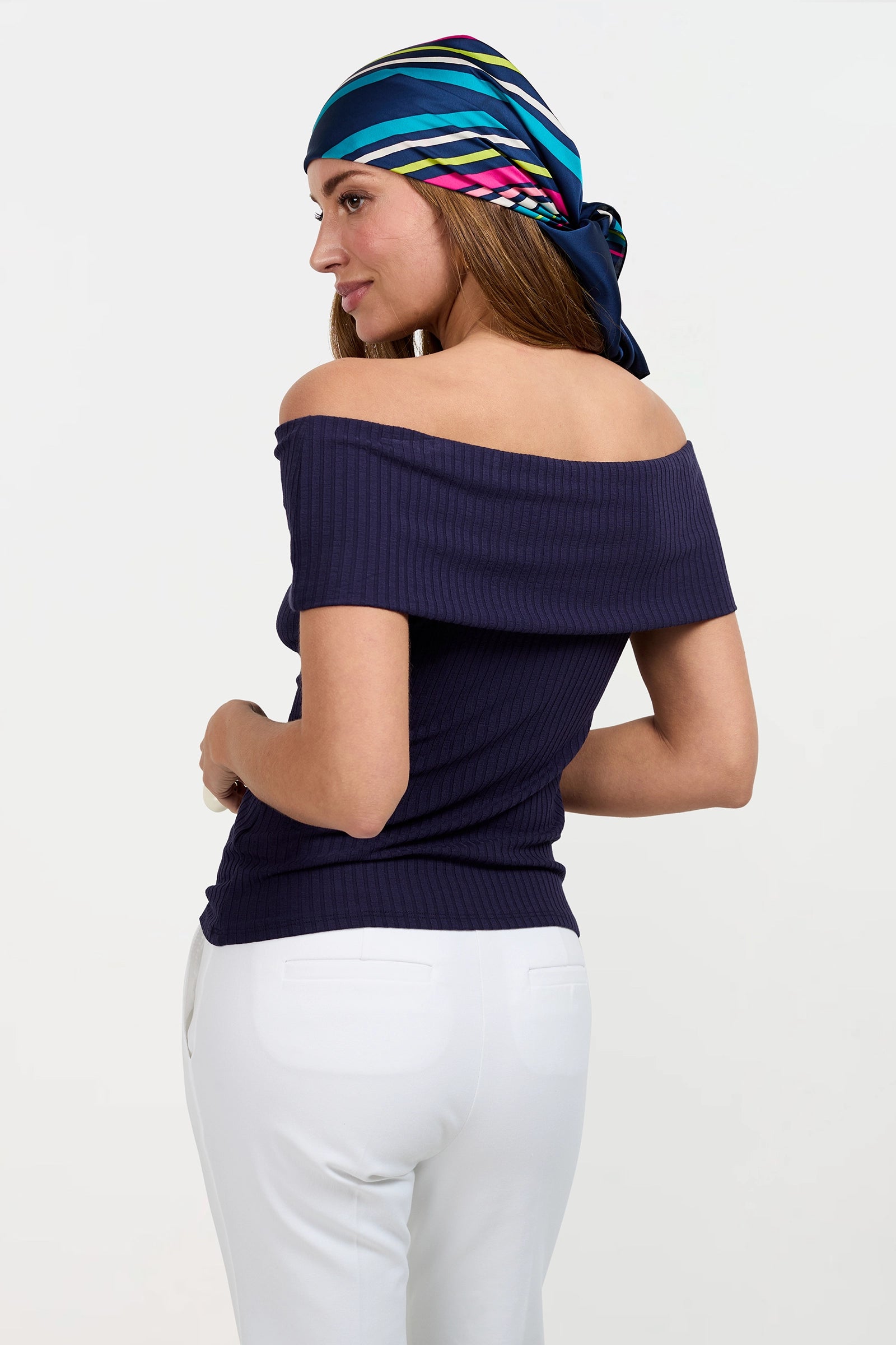 A woman in the Prizma Off-Shoulder Top (navy), white pants, and a colorful striped headscarf stands turned sideways, looking over her shoulder against a plain light background.