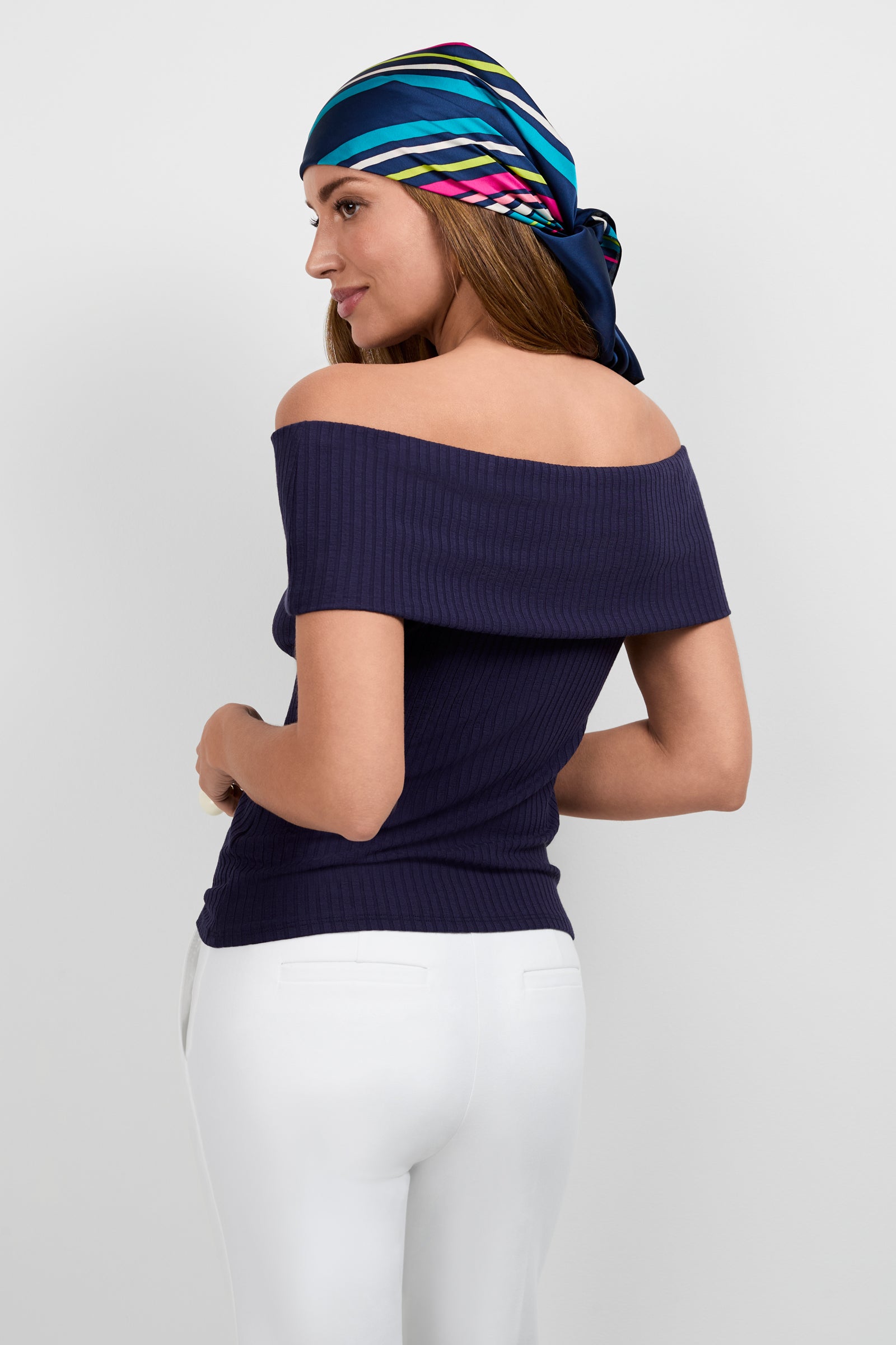 A woman models the Prizma Off-Shoulder Top in navy with white pants and a colorful striped headscarf, standing slightly turned and looking over her shoulder against a plain background.