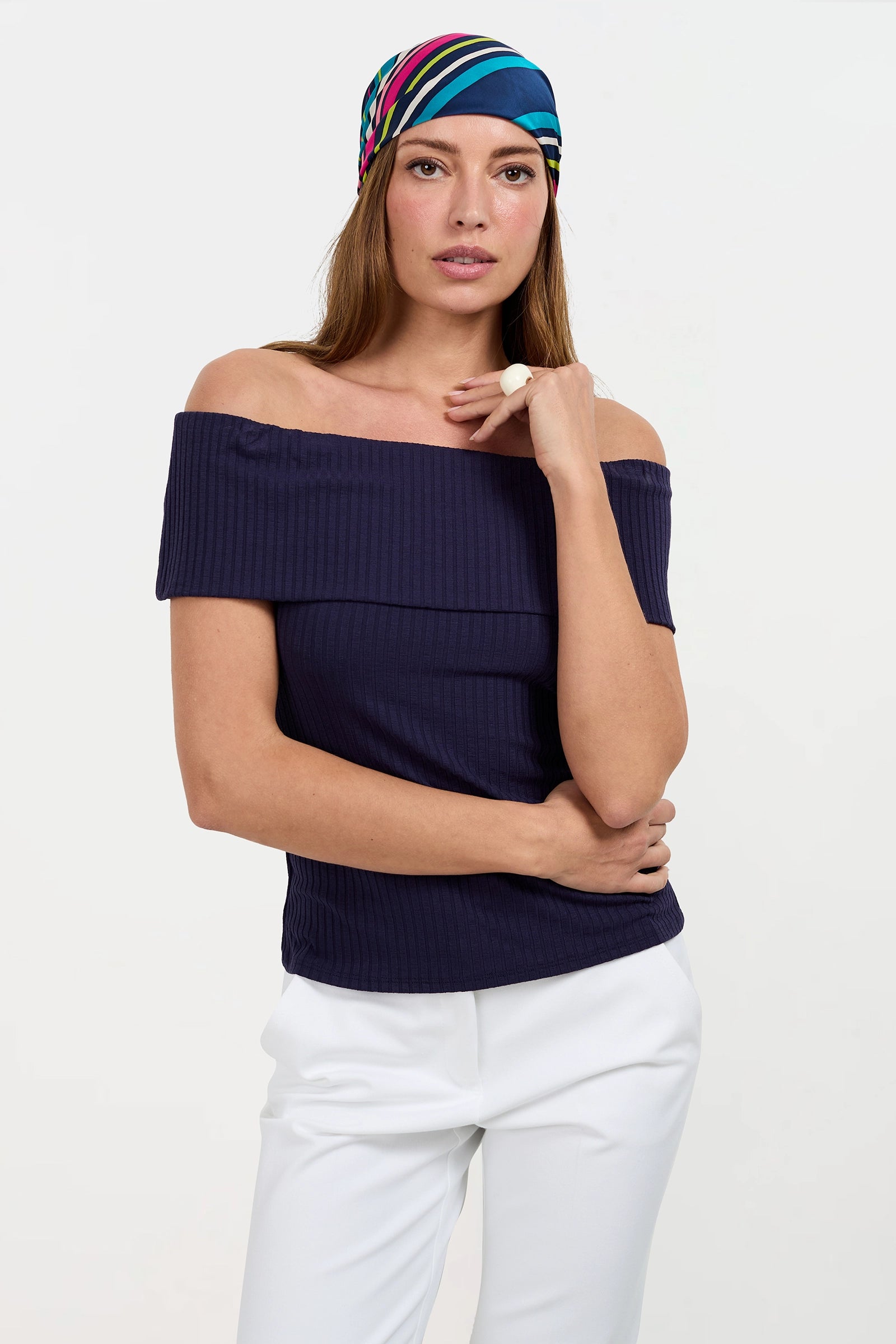 A woman models the Prizma Off-Shoulder Top in navy, paired with white pants and a colorful striped headscarf, posing with one hand on her hip and the other touching her shoulder against a plain white background.