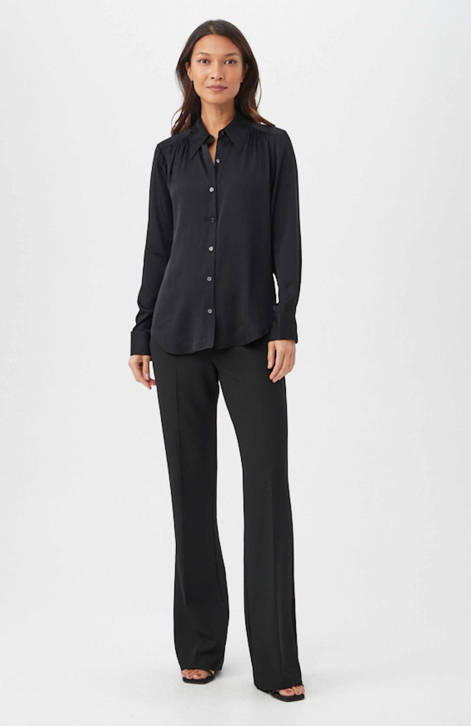 A woman with long hair and a neutral expression showcases the Cassavetes Long-Sleeve Stretch Silk Button-Up Shirt, paired with straight-leg trousers and open-toe black shoes against a plain white background.