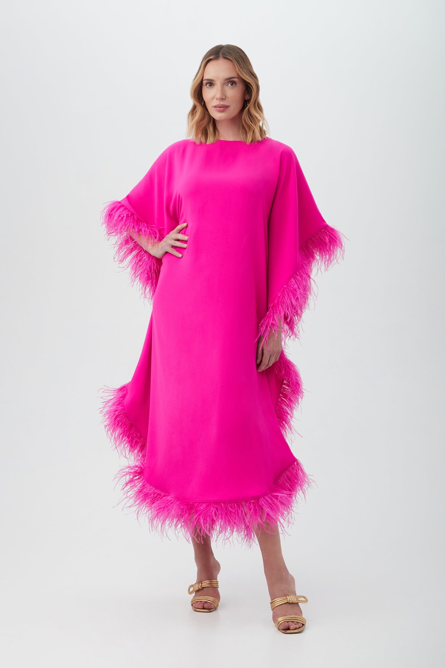 A woman poses against a light gray background wearing the Neena Maxi Caftan Dress in Trina Pink, featuring a loose fit and feather trim on the sleeves and hem, paired with gold open-toe sandals.