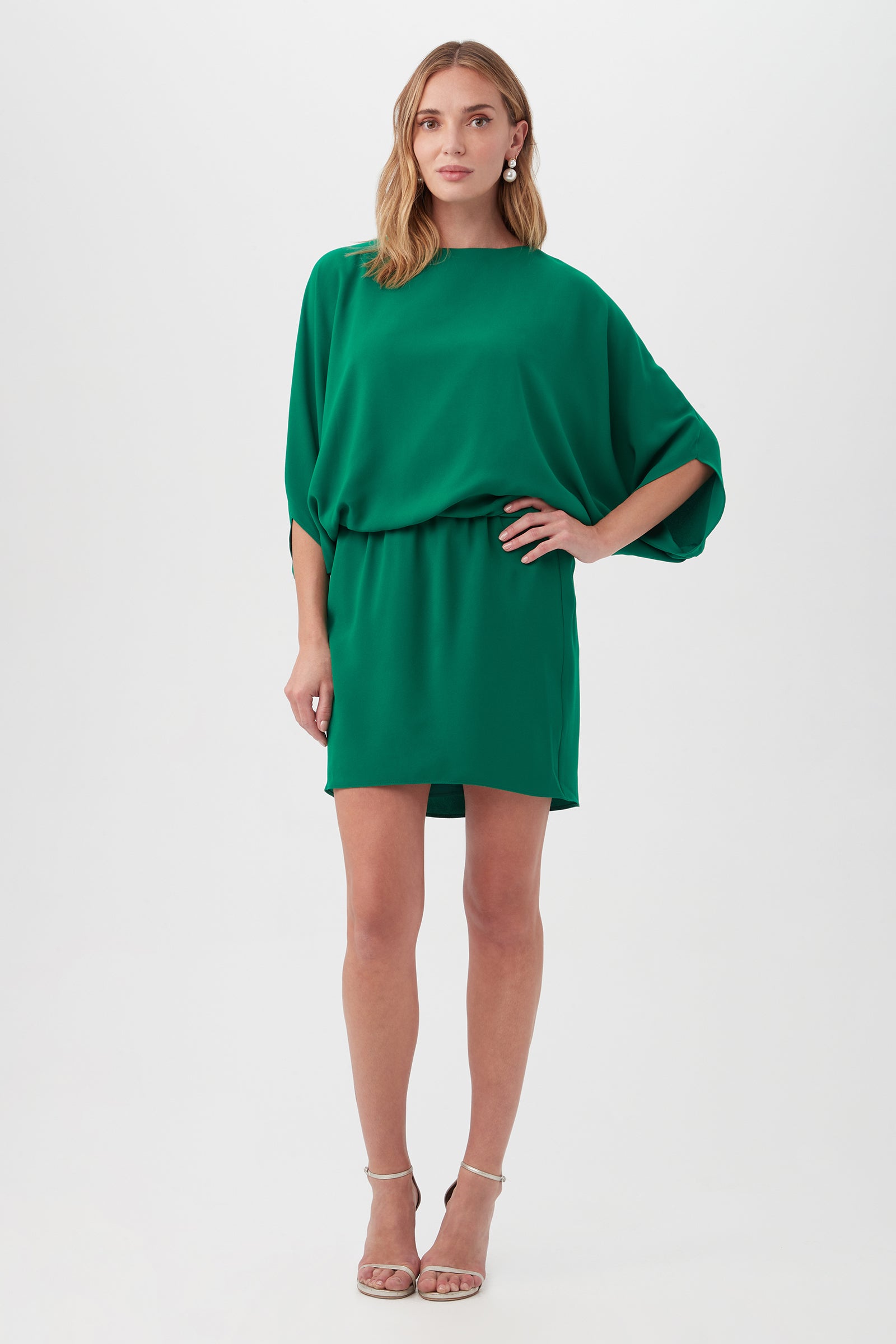 Woman stands against a white background wearing the Manhattan Dressâ€”a loose, knee-length green dress with batwing sleeves and belted waistâ€”paired with nude strappy high-heeled sandals.
