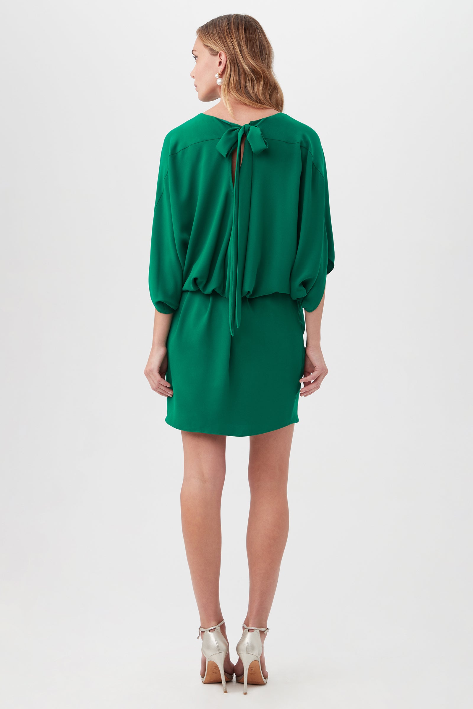 A woman faces away in the Manhattan Dress, a loose-fitting, knee-length green dress with a tied neck bow. She has wavy light brown hair and wears white high-heeled sandals against a plain white background.