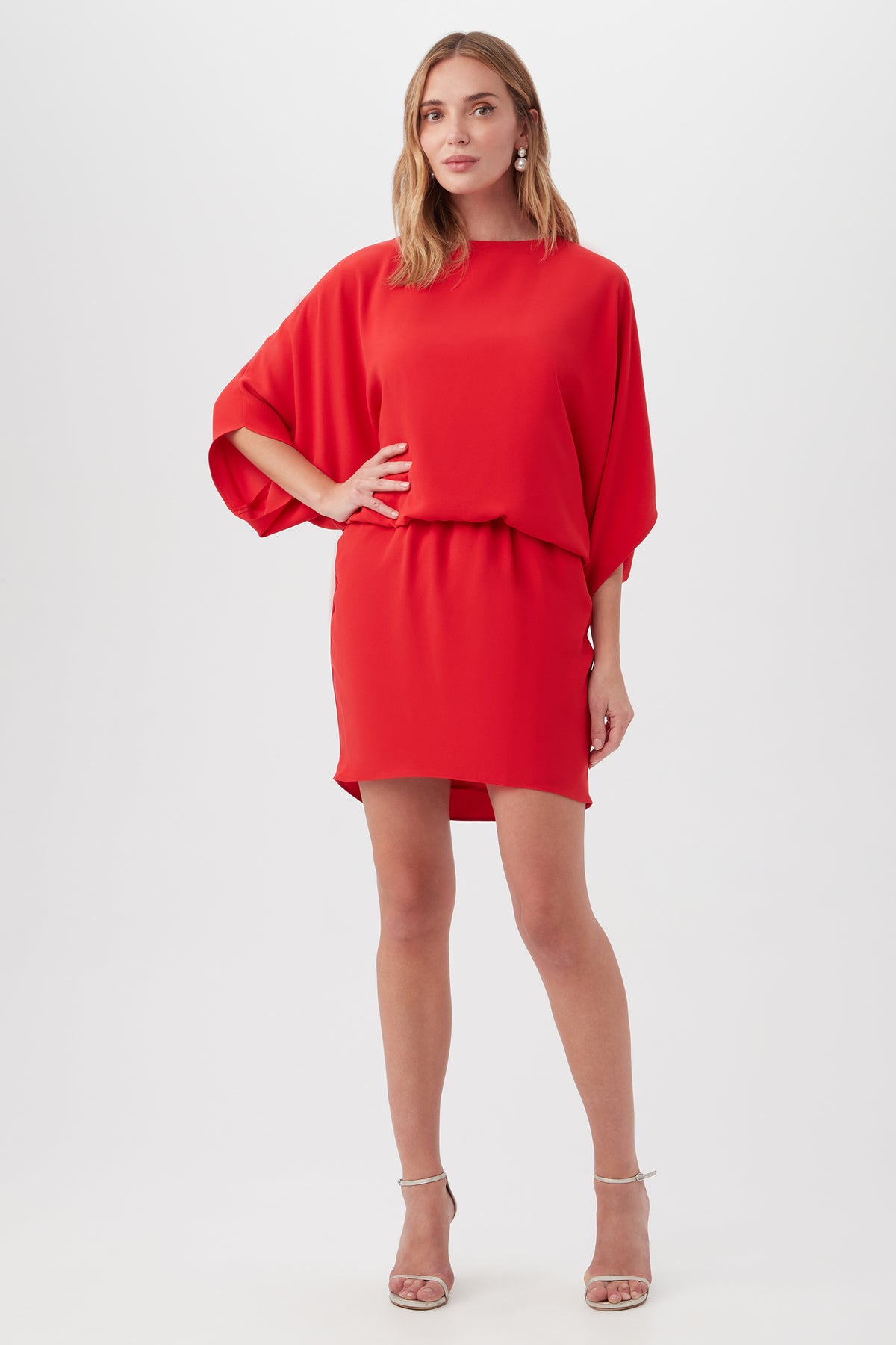 A woman stands confidently in the Manhattan Dressâ€”a vibrant red, loose-fit, short-sleeve piece with a relaxed waist and short hemâ€”paired with strappy nude high-heeled sandals, set against a plain light background.
